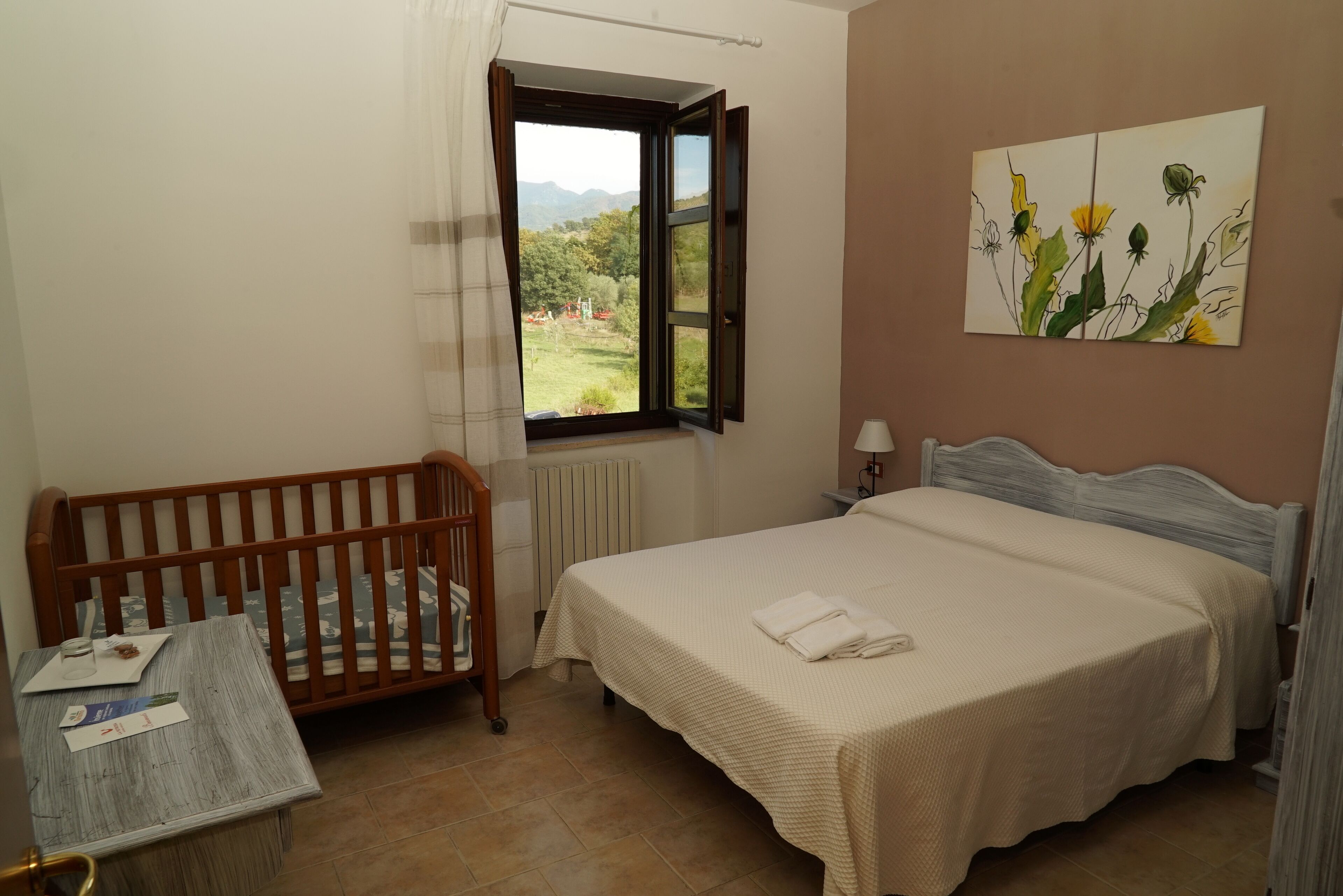 A bedroom with a wooden crib, a bed with a white bedspread, a window with a view of greenery, and a painting on the wall.