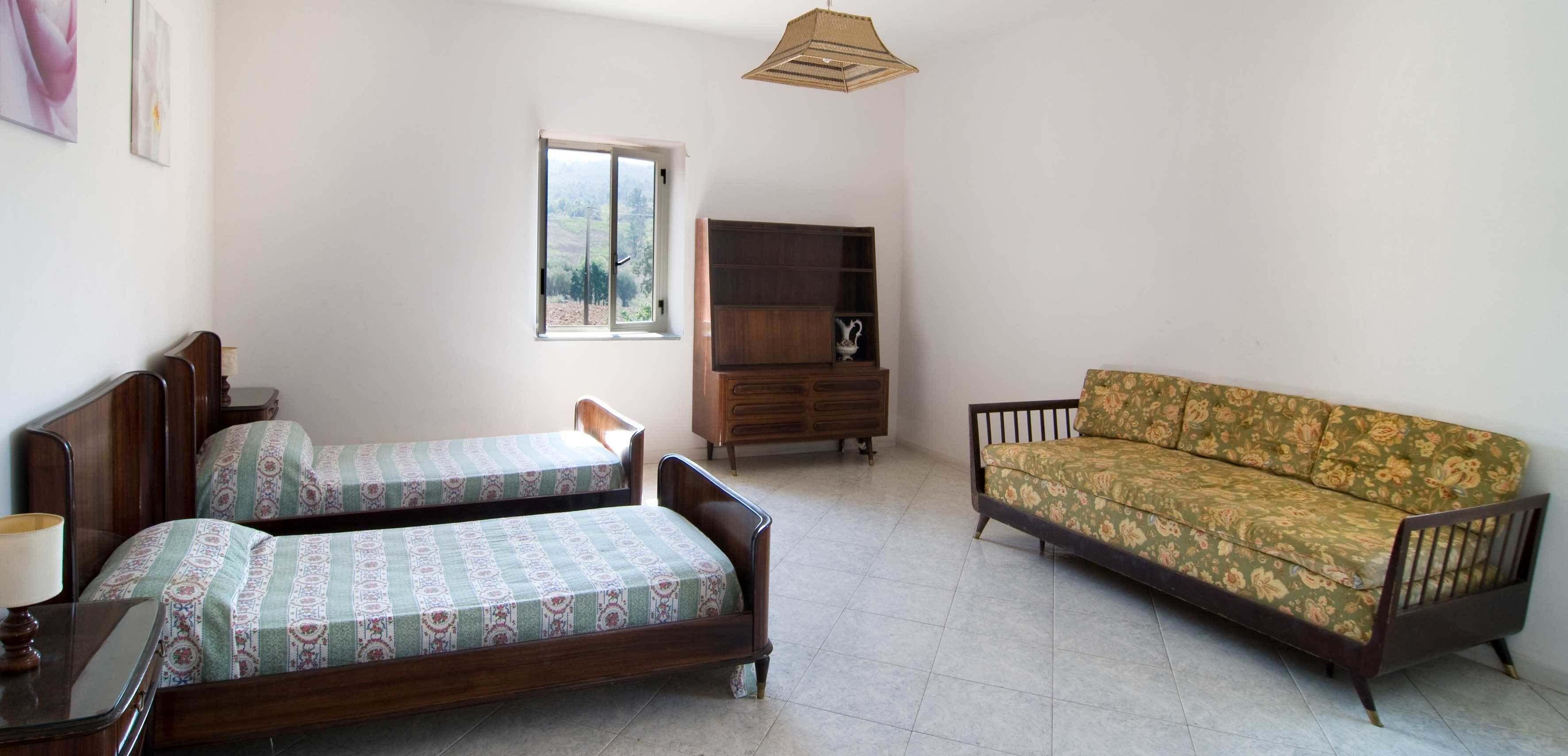 A bedroom with two wooden beds, a sofa, a wooden cabinet, and a window with a view of trees.