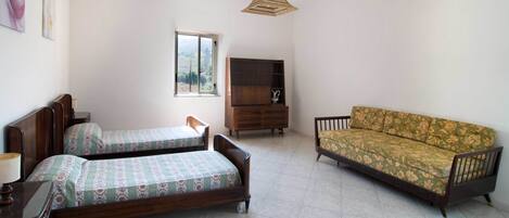 A bedroom with two wooden beds, a sofa, a wooden cabinet, and a window with a view of trees.