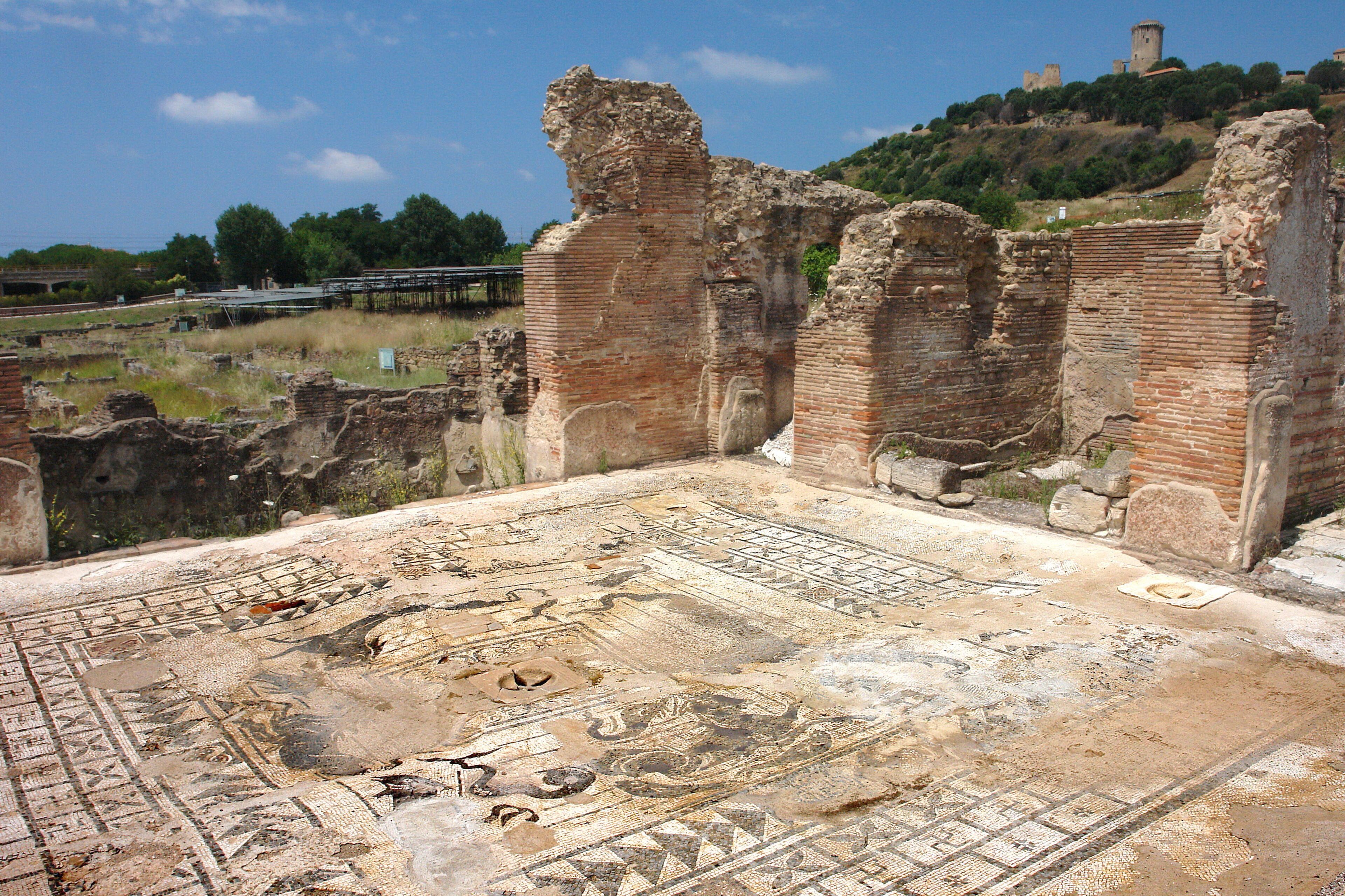 Ancient ruins with a mosaic floor, brick walls, and a hill with a tower in the background.