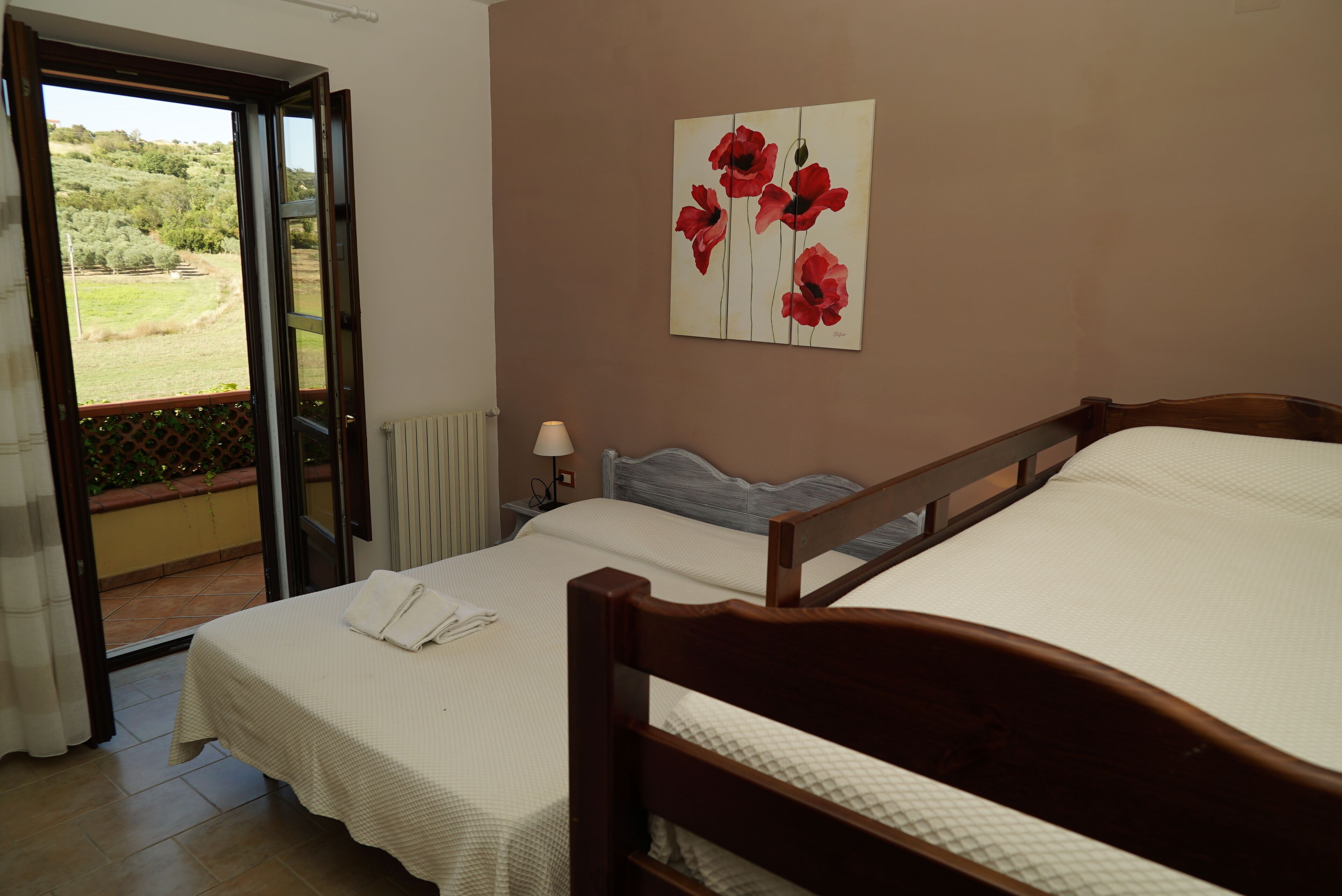 Un dormitorio con dos camas, vistas a un jardín por una puerta abierta, un cuadro de flores rojas en la pared y una lámpara de noche.