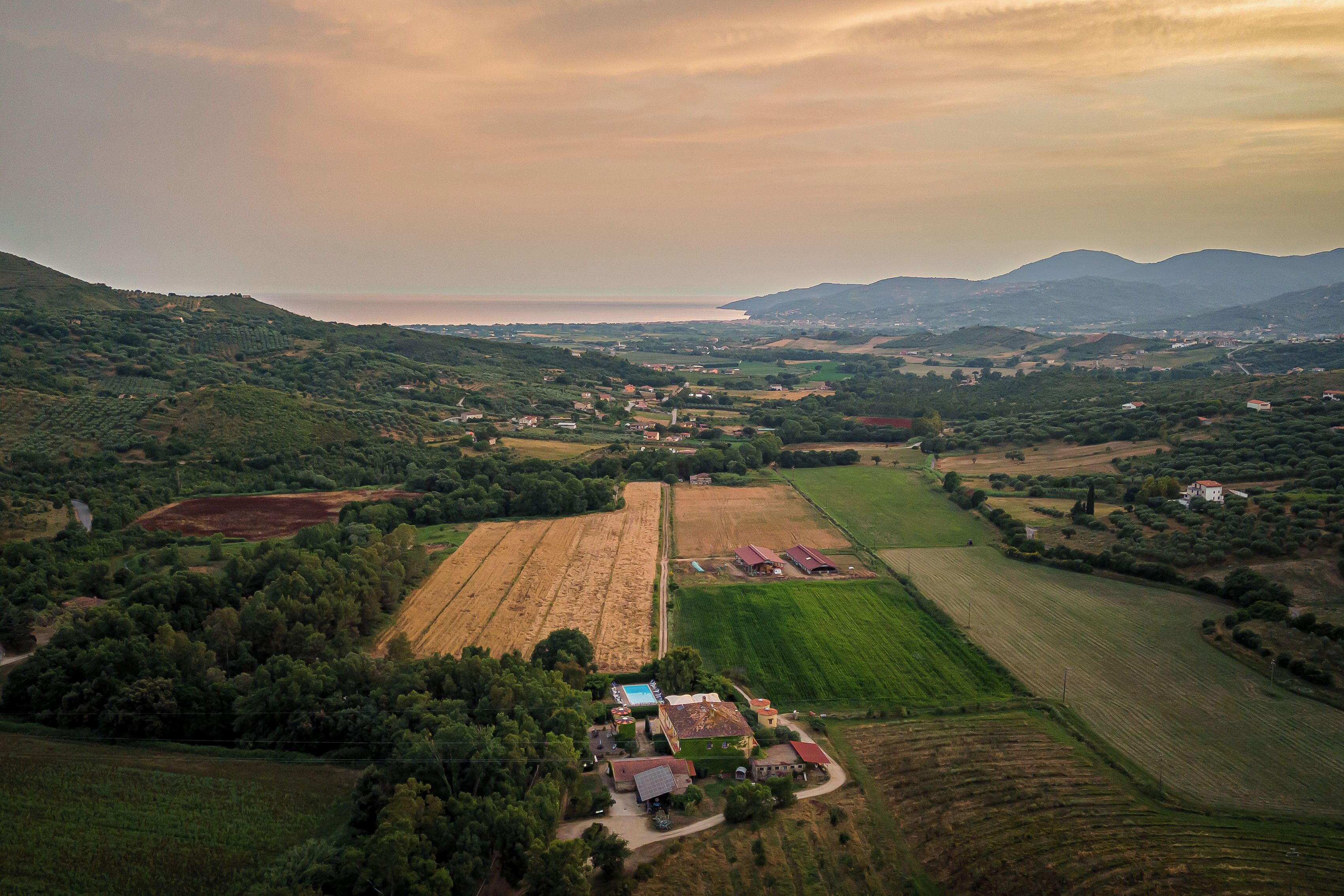 Vista aerea di un paesaggio rurale con campi, un piccolo gruppo di edifici e montagne in lontananza.
