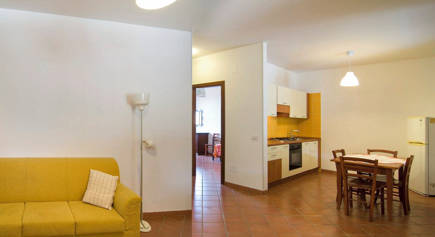 A compact living space with a sofa, a dining table with chairs, a kitchen area with cabinets and a refrigerator, and a floor lamp.