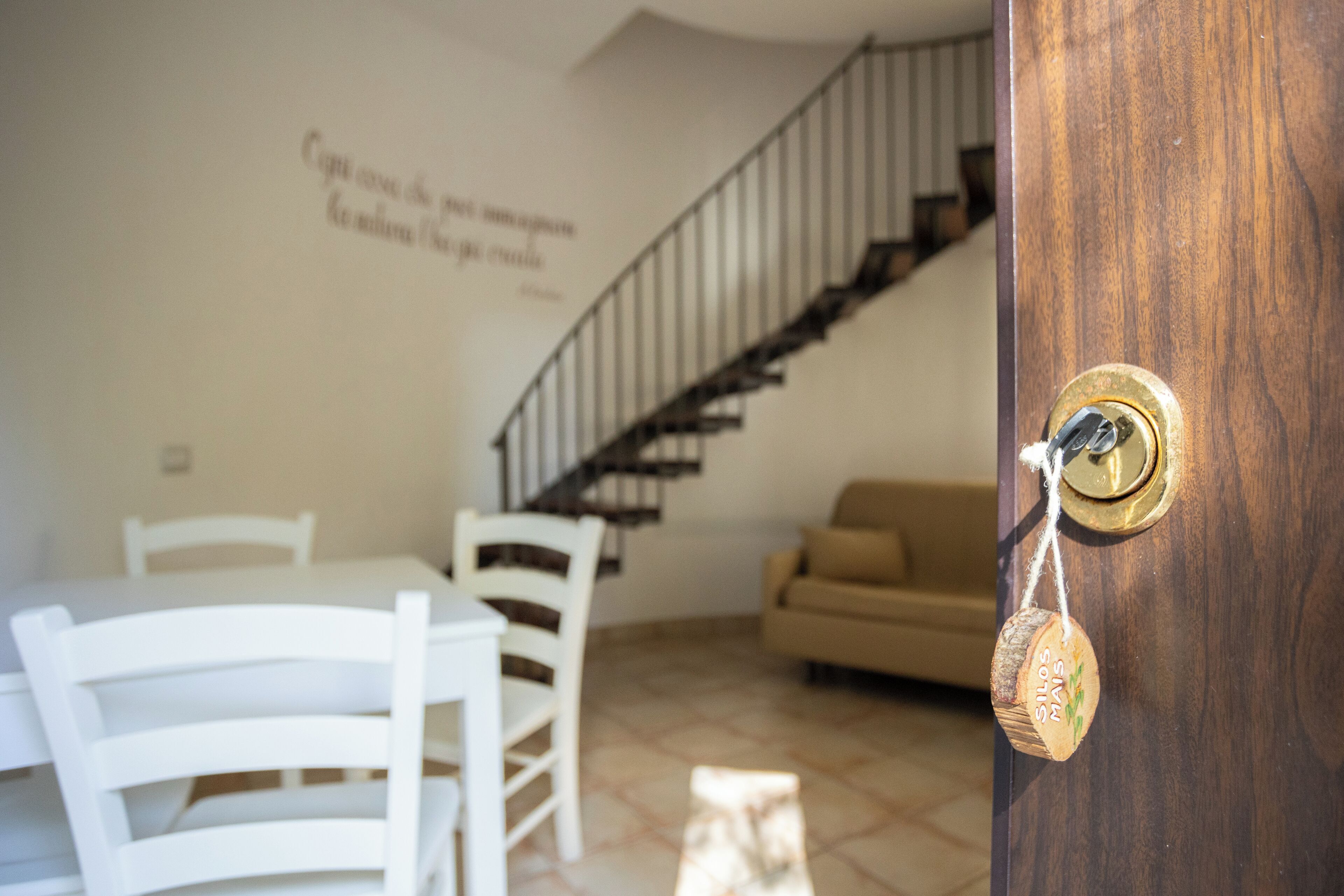 Un coin repas avec une table et des chaises blanches, un escalier en arrière-plan et une porte en bois dotée d’une poignée de porte en laiton.