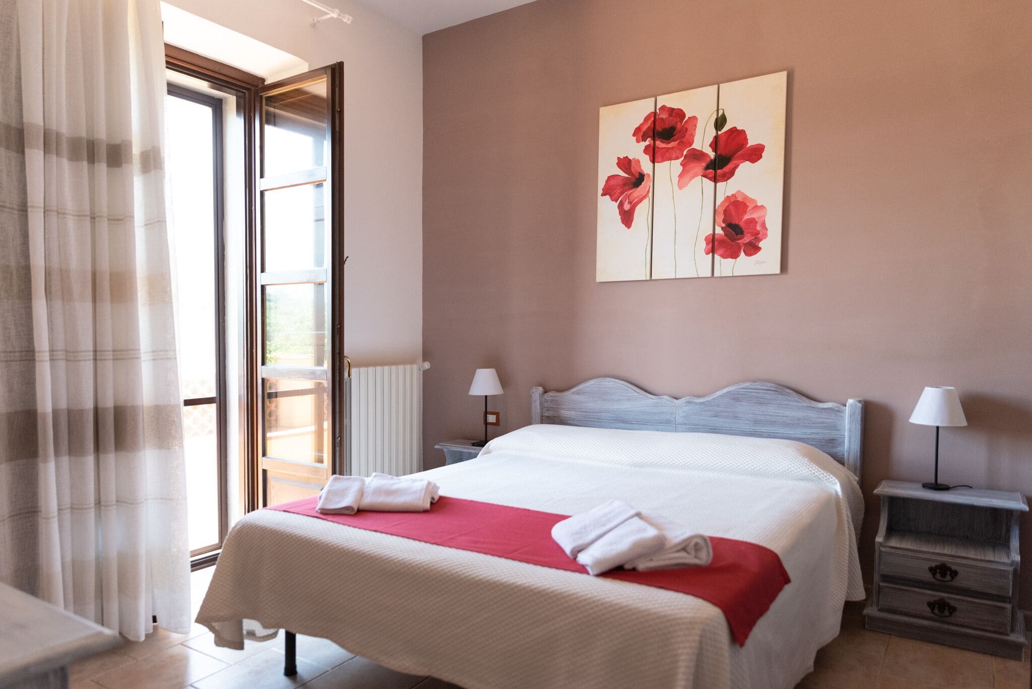 A hotel room with a bed, bedside tables, a window with curtains, and a wall with a floral painting.
