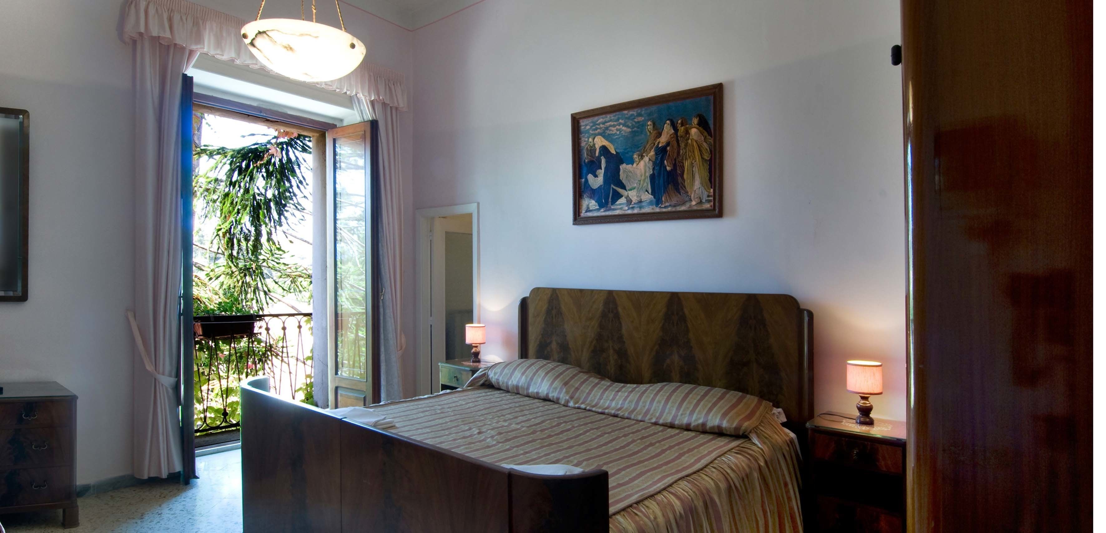 A bedroom with a bed, a nightstand, a lamp, a painting, and a window with curtains.
