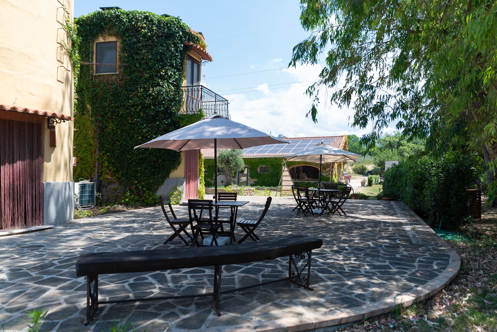 Une terrasse en pierre avec des sièges extérieurs, entourée de verdure et bordée d’un bâtiment doté d’un balcon.