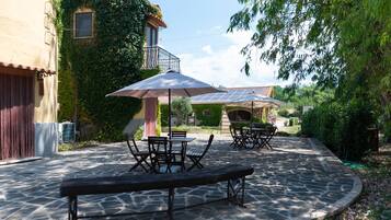 Une terrasse en pierre avec des sièges extérieurs, entourée de verdure et bordée d’un bâtiment doté d’un balcon.