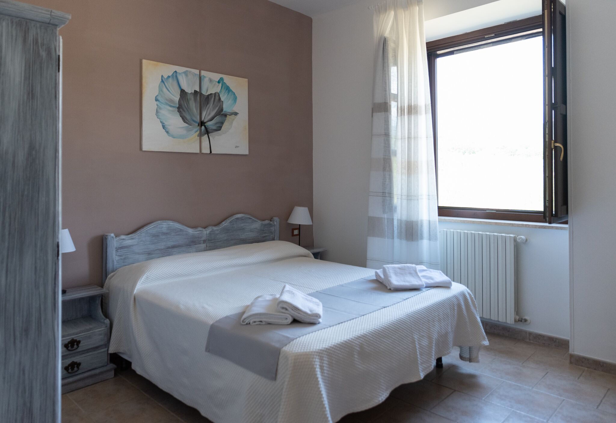 A neatly made bed with white linens and towels, a painting on the wall, and a window with curtains.