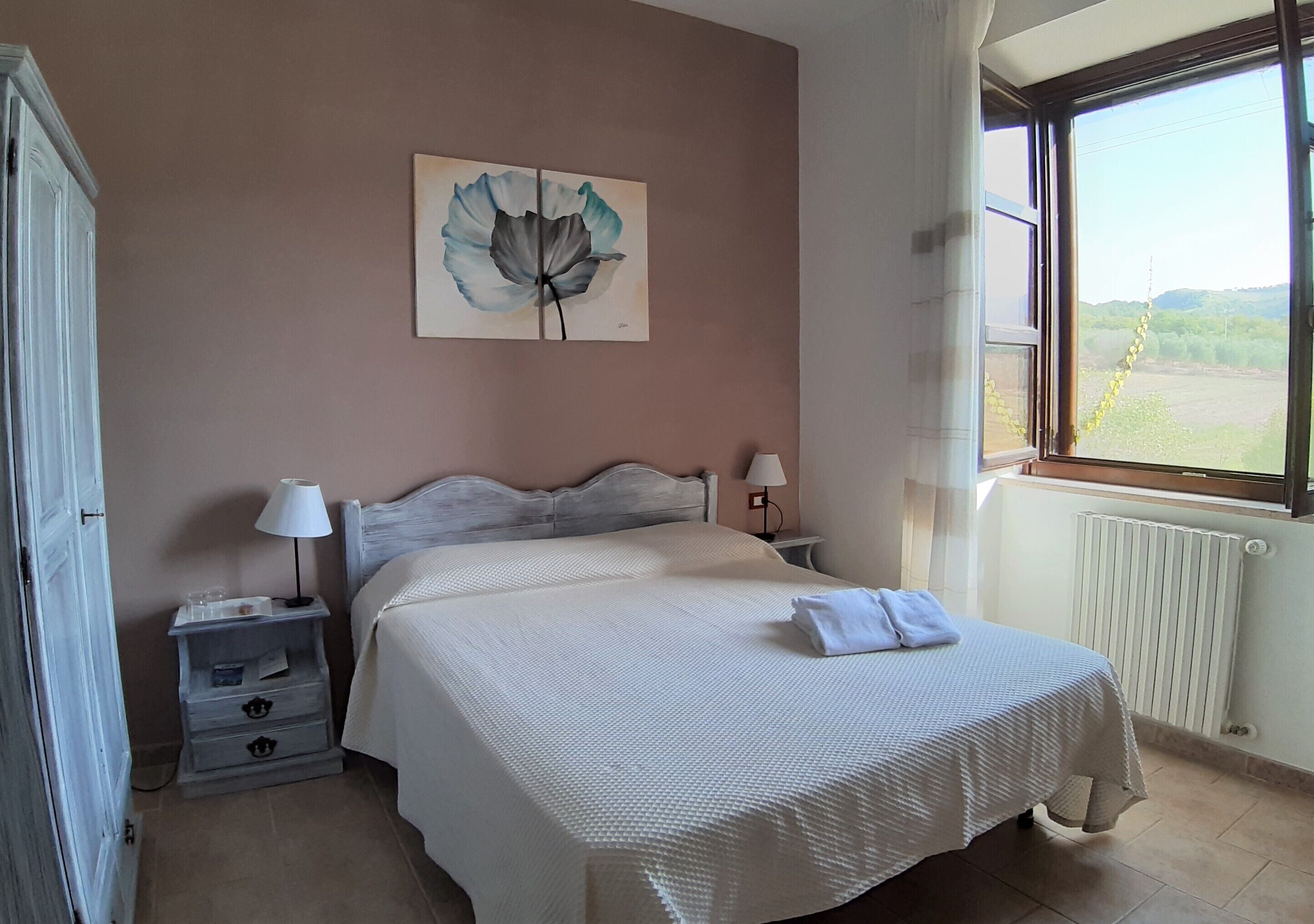 A bedroom with a bed, bedside tables, a lamp, a radiator, and a window with a view of the countryside.