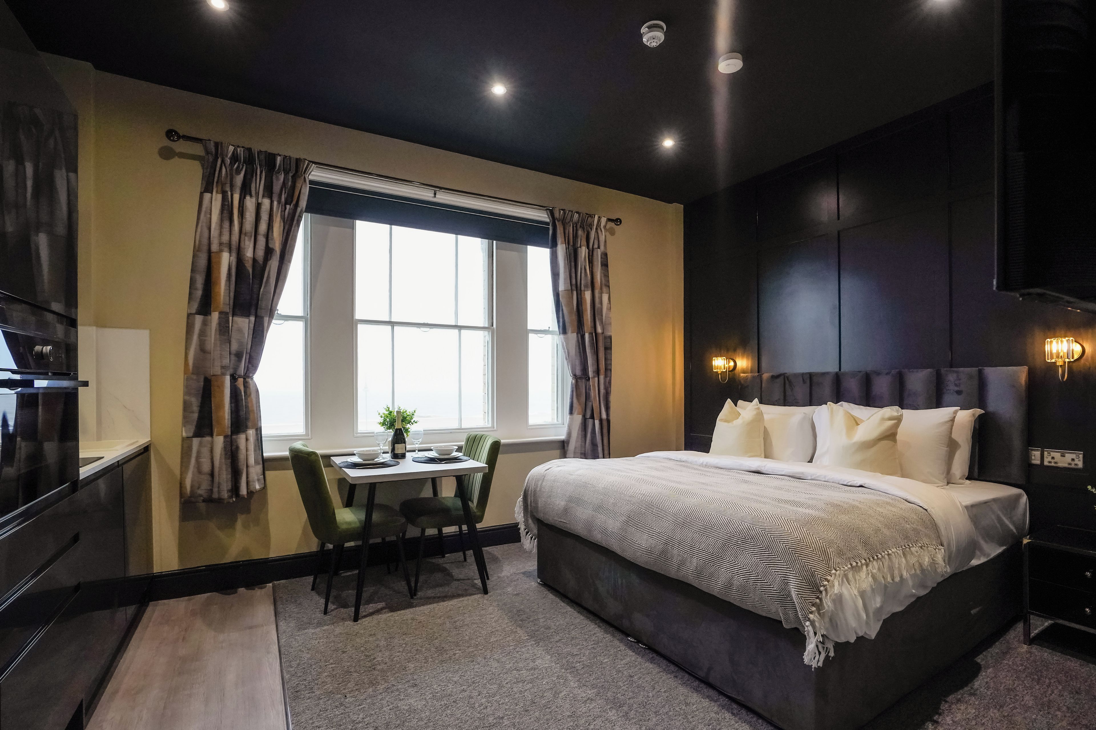 Ein modernes Schlafzimmer mit einem großen Bett, einem Esstisch mit Stühlen und Blick ins Freie durch das Fenster.