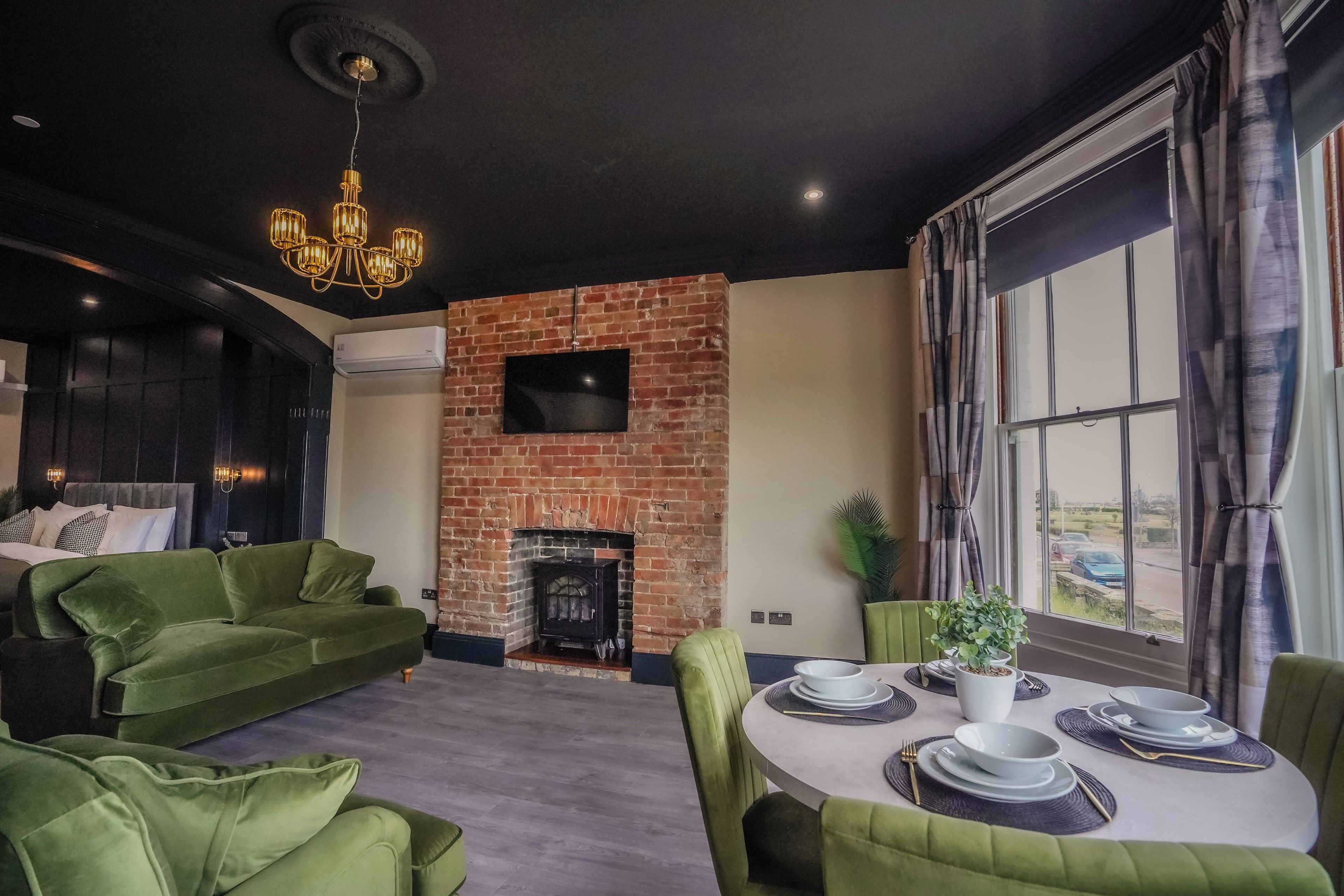 Una sala de estar moderna con chimenea de ladrillo, sofás verdes, una mesa de comedor preparada para cuatro personas y un ventanal con cortinas.