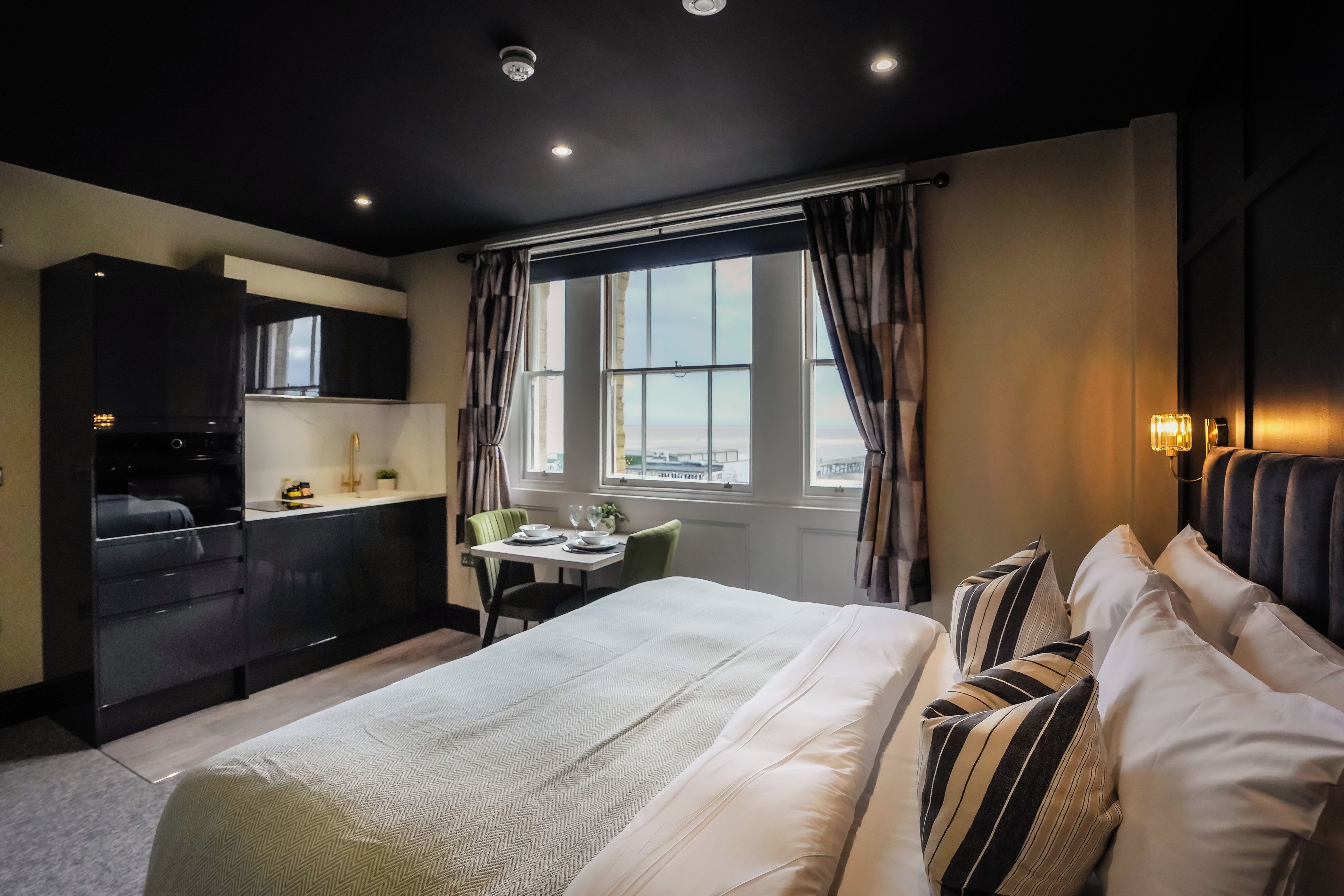 A hotel room with a bed, a dining table, a kitchen area, and a view of the city.