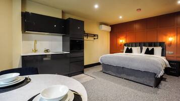 Habitación de hotel con cama, mesitas de noche, una pequeña cocina con fregadero y armarios, y una mesa de comedor con platos y cubiertos.