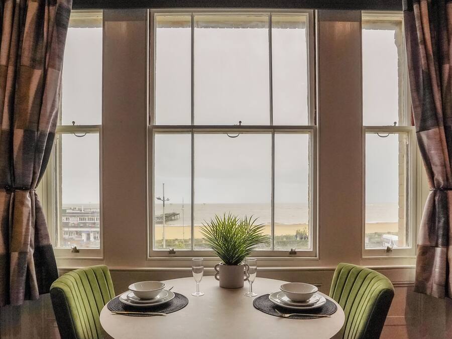 A dining area with a table set for two, a potted plant, and a view of the sea through large windows.