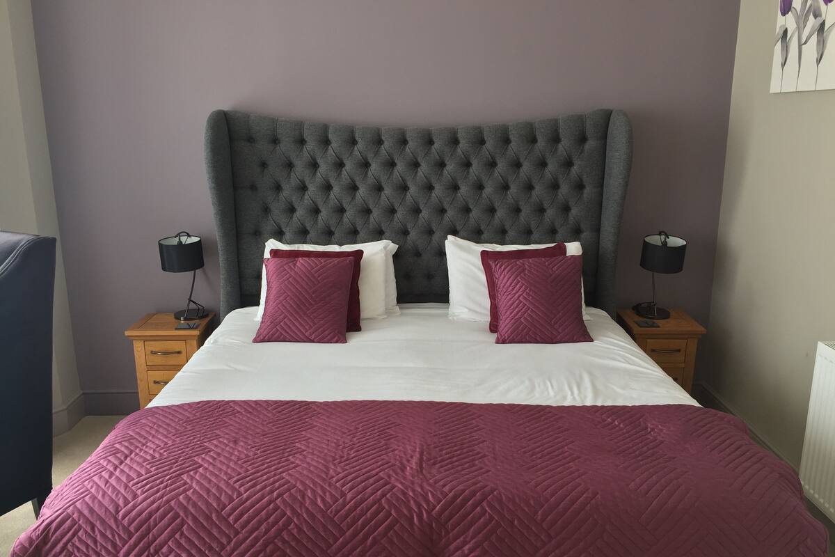 A neatly made bed with a grey tufted headboard, two bedside tables with lamps, and a purple quilted bedspread.