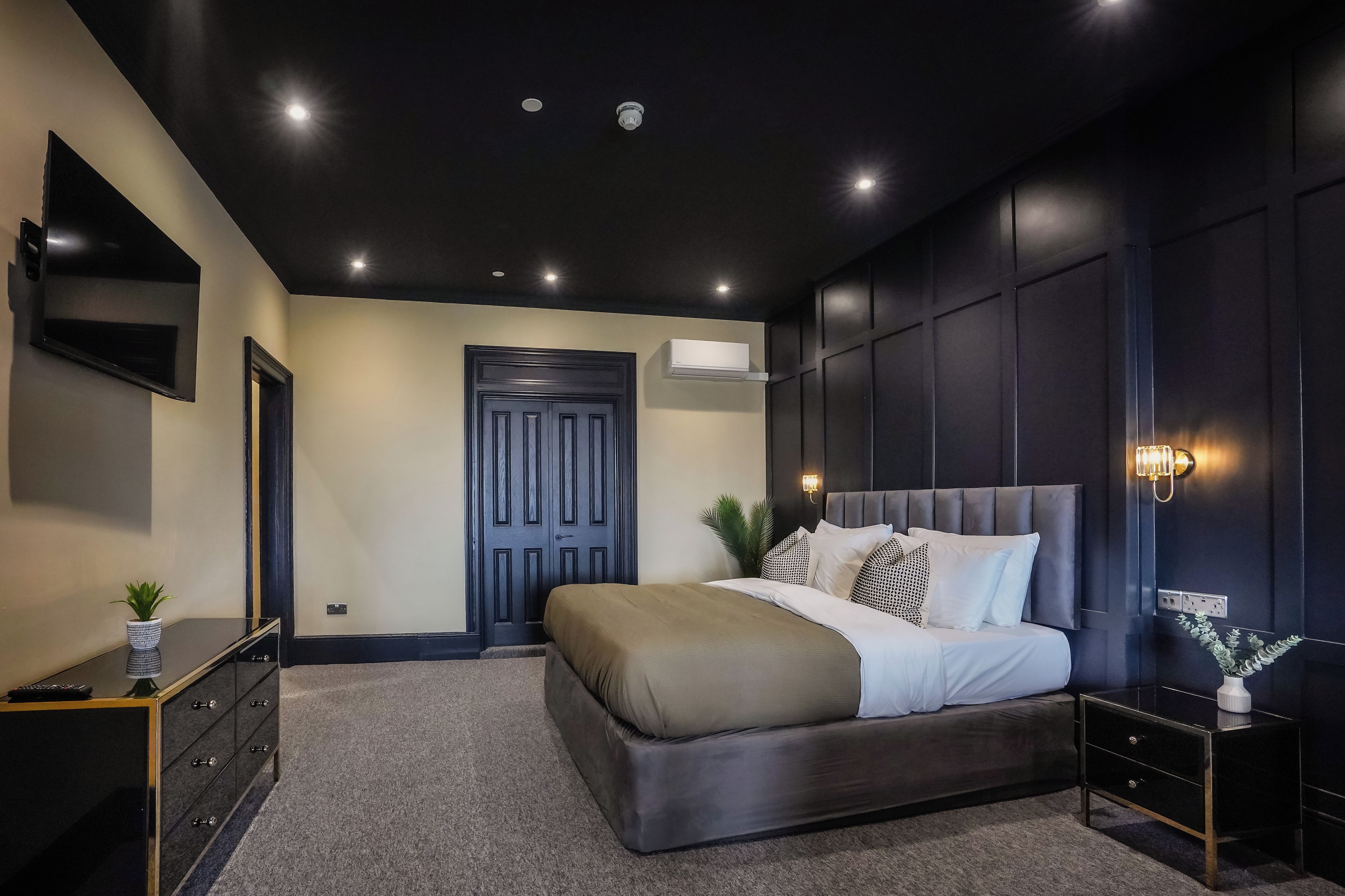 Ein modernes Schlafzimmer mit einem großen Bett, einem Schminktisch, einem an der Wand montierten Fernseher und einer Tür.