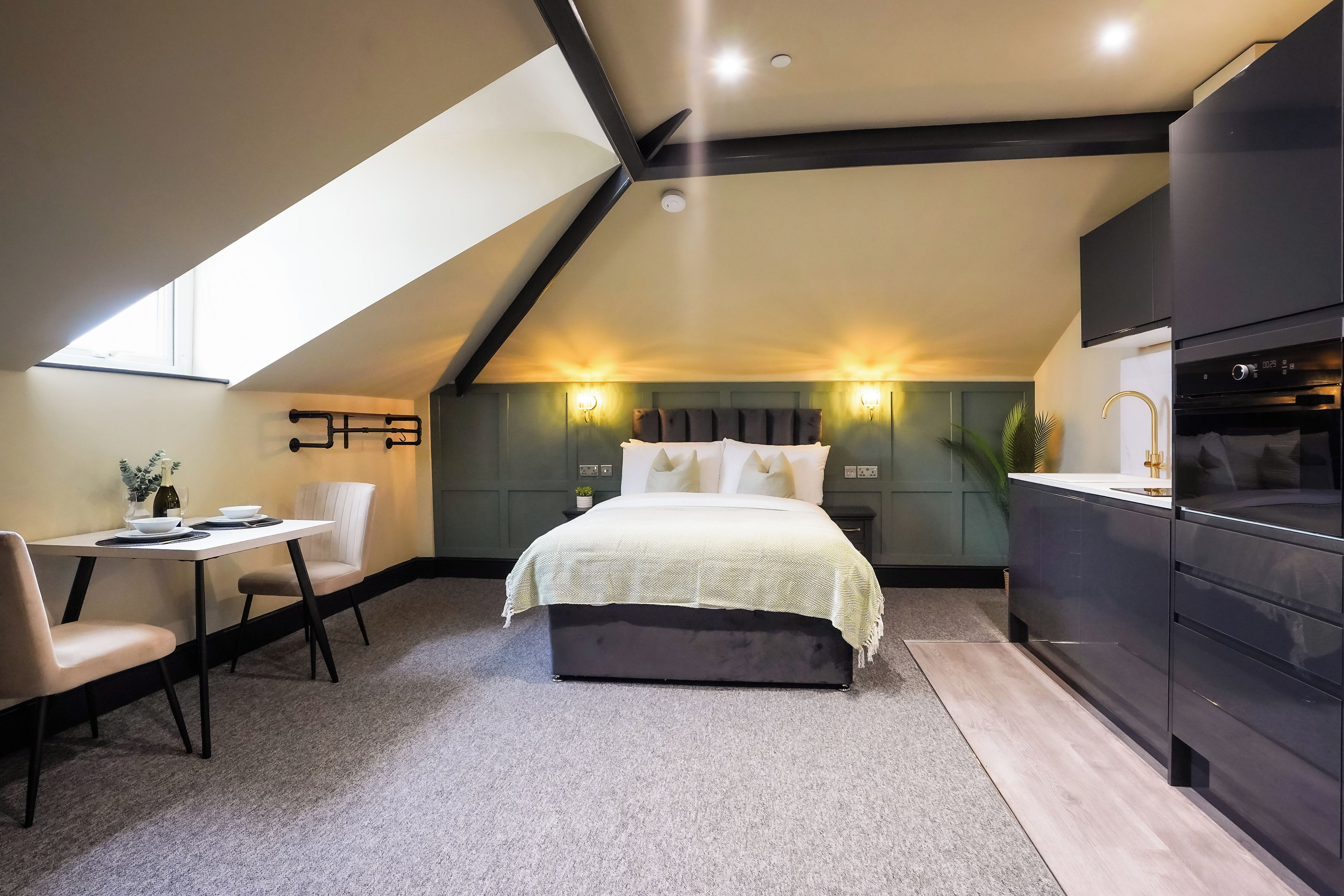 A modern bedroom with a sloped ceiling, a bed with a herringbone-patterned bedspread, a desk with a chair, and a built-in kitchen area with a stove.