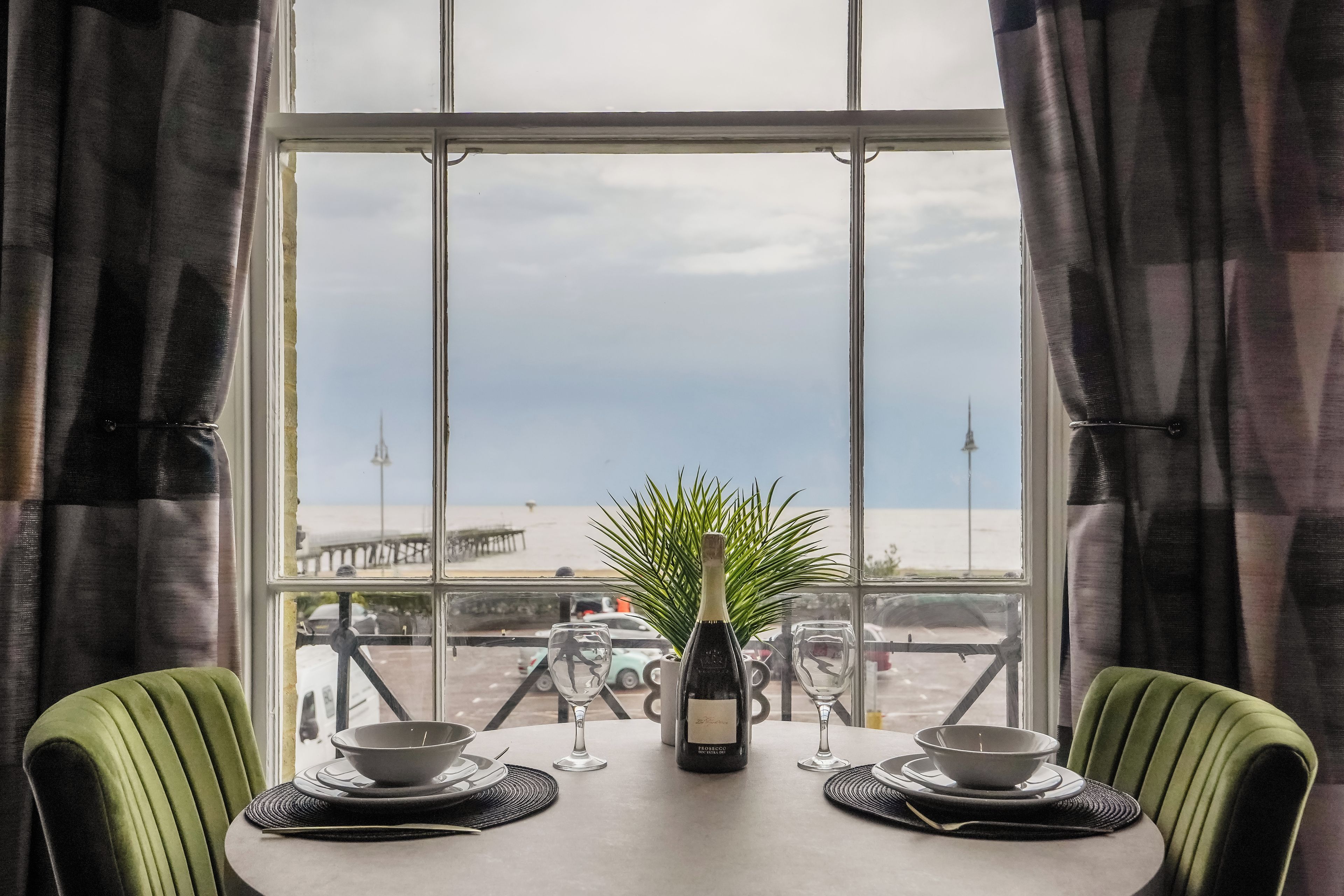 A dining table with a bottle and glasses, overlooking a beach and pier.