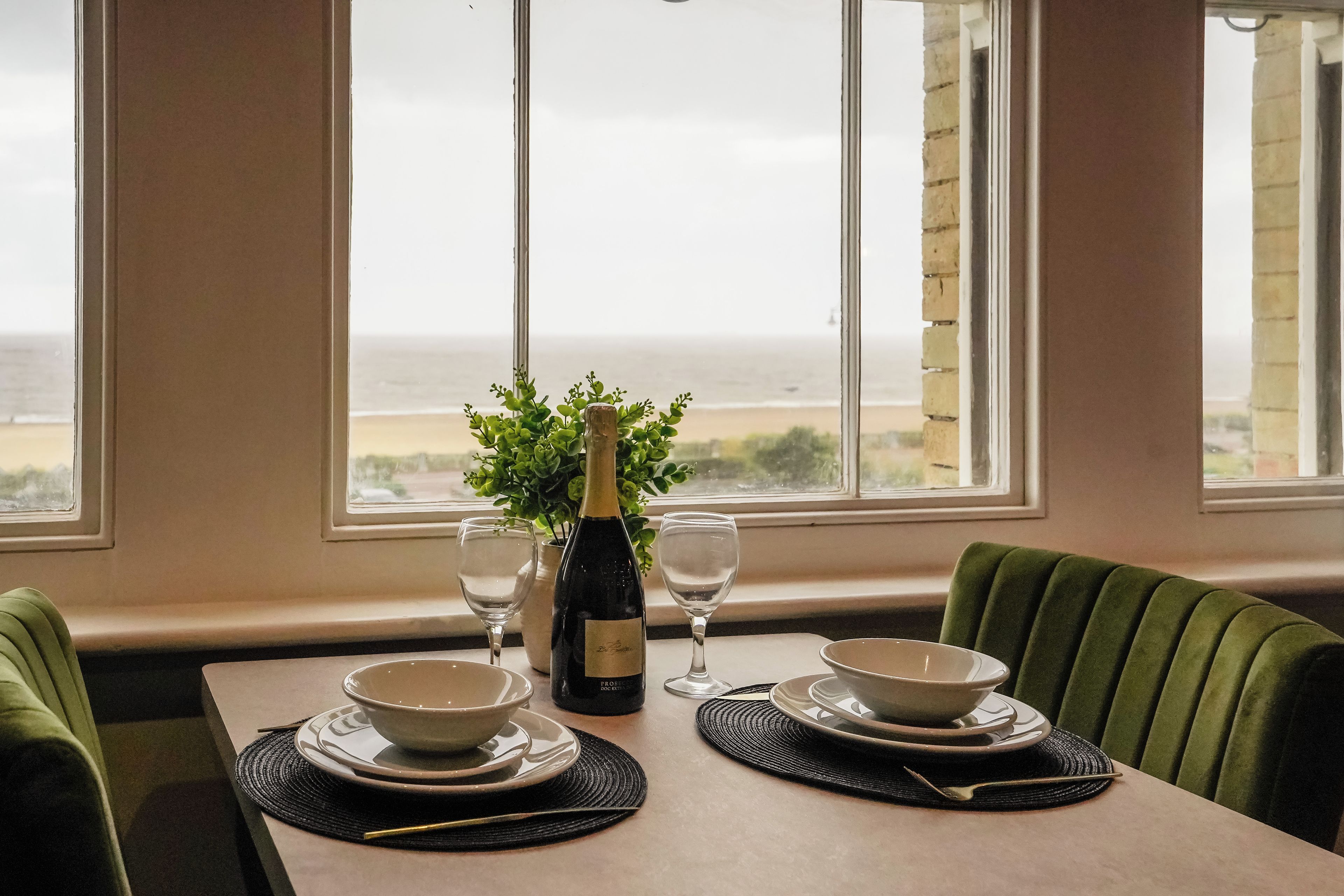 A table set for dining with a view of the sea, a bottle of wine, and a green plant.