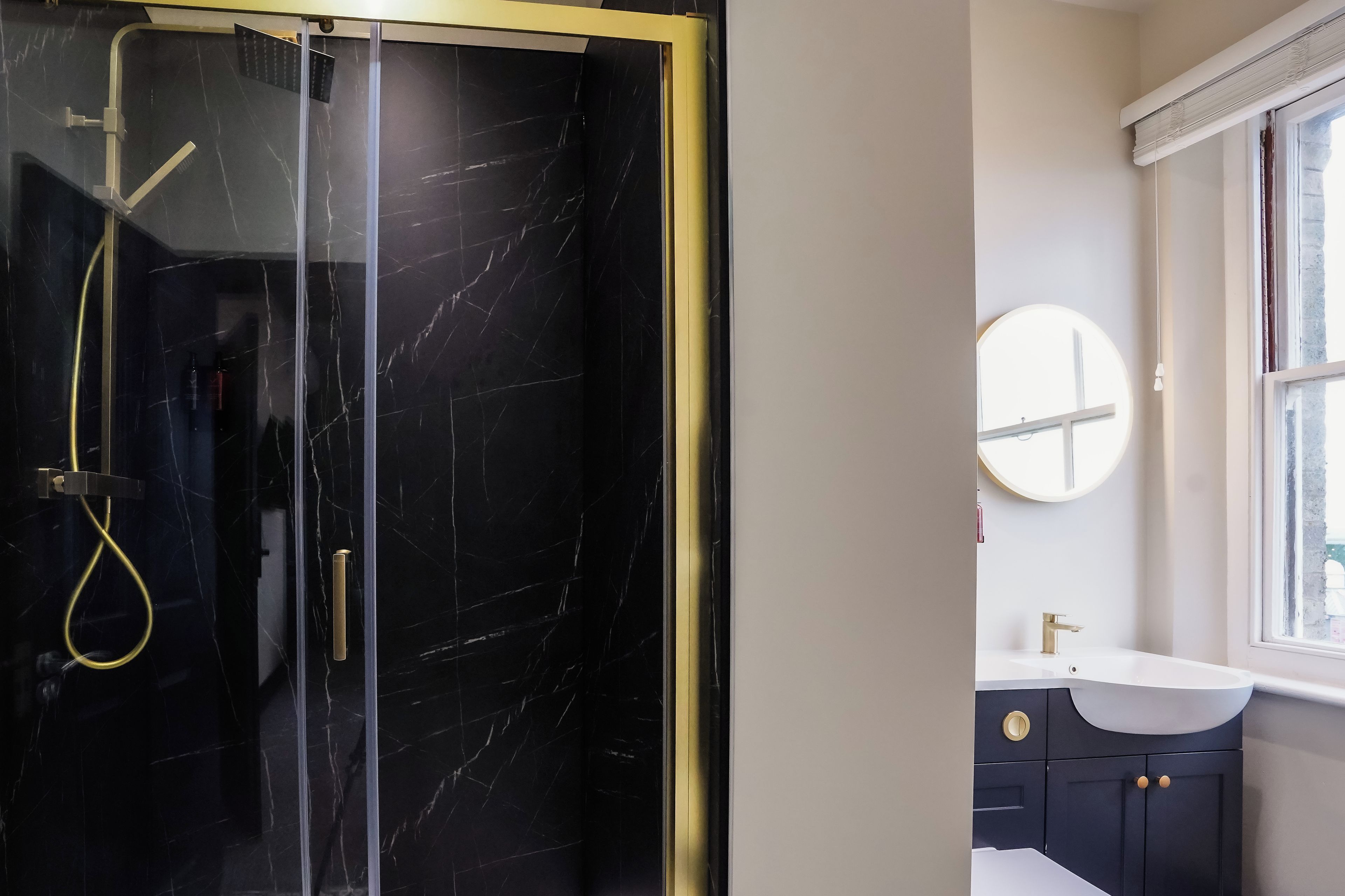 A modern bathroom with a glass-enclosed shower, a round mirror, and a dark-colored vanity with gold handles.