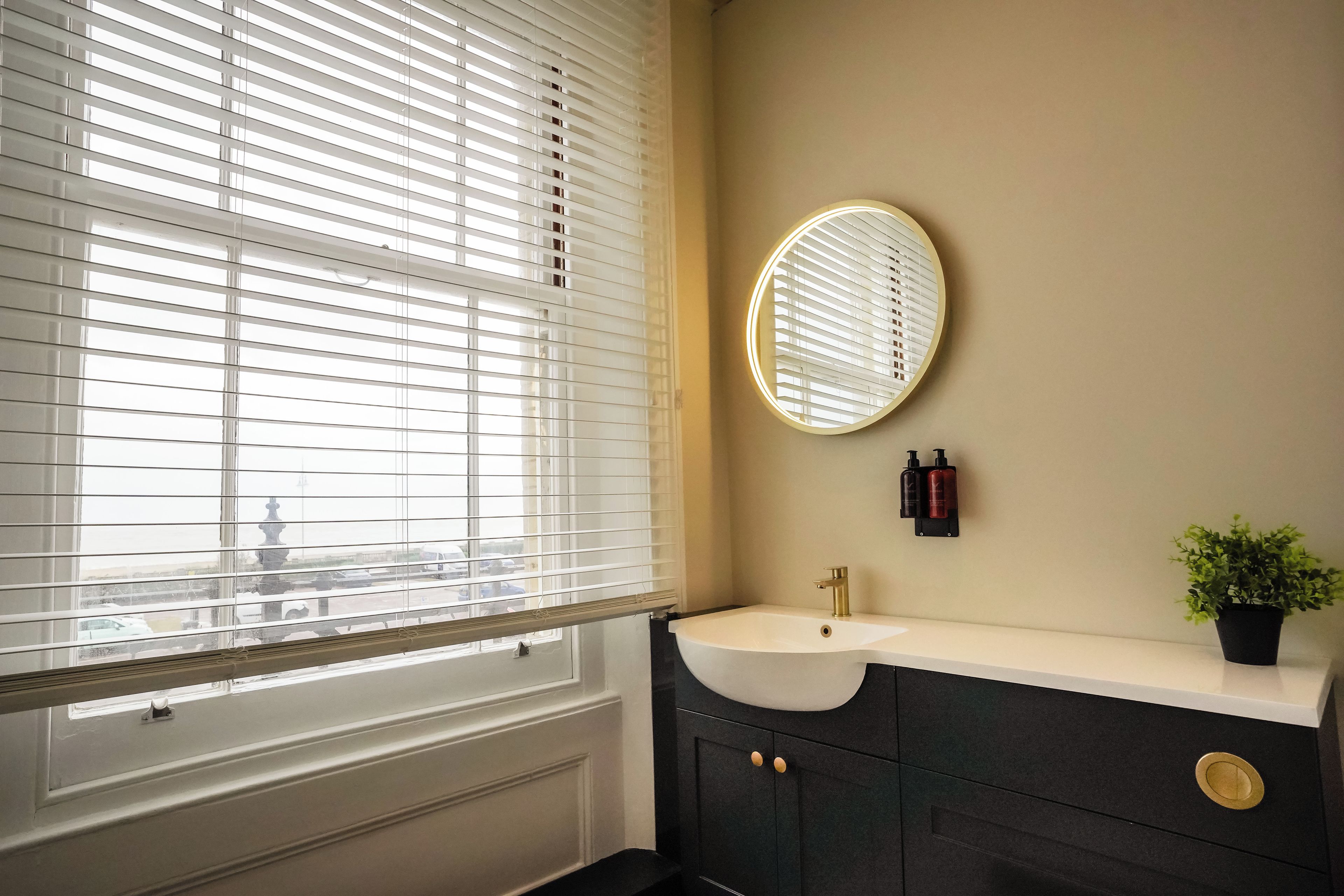A bathroom with a white sink, dark cabinets, a round mirror, and a potted plant.