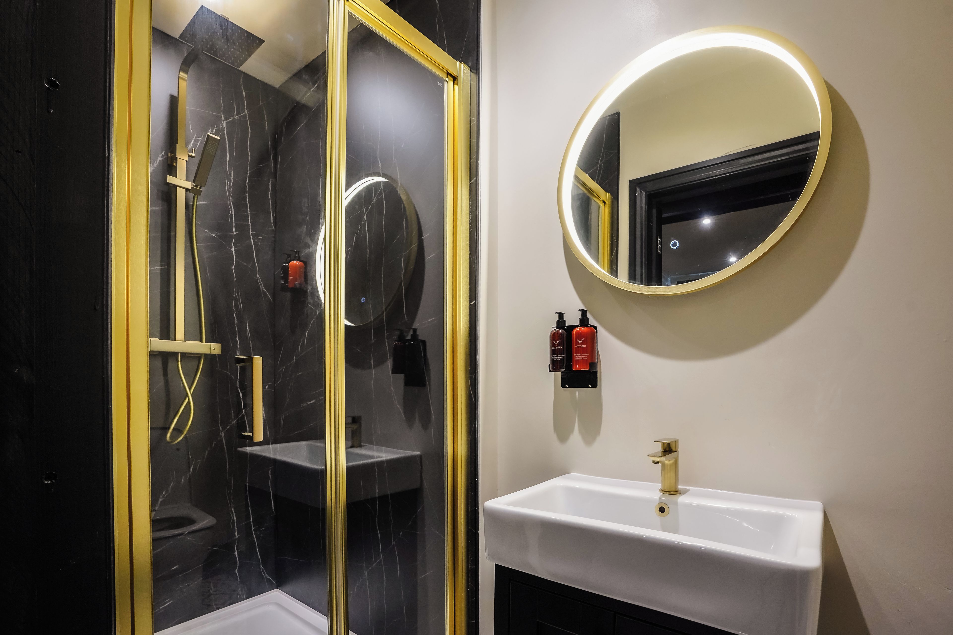 A bathroom with a glass-enclosed shower, a white sink, and a round mirror with a gold frame.