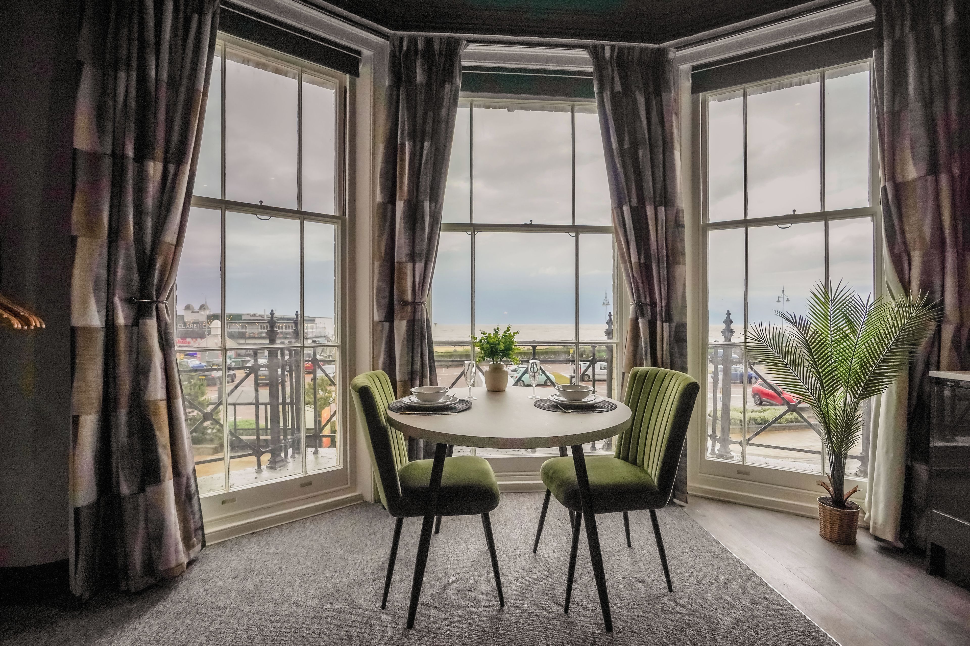 Habitación de esquina con una mesa redonda, dos sillas, una planta en maceta y vistas de la ciudad a través de amplios ventanales.