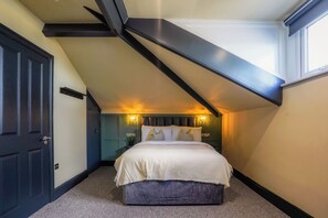Un dormitorio con techo inclinado, una cama con ropa de cama blanca, una mesita de noche con lámpara y una puerta.