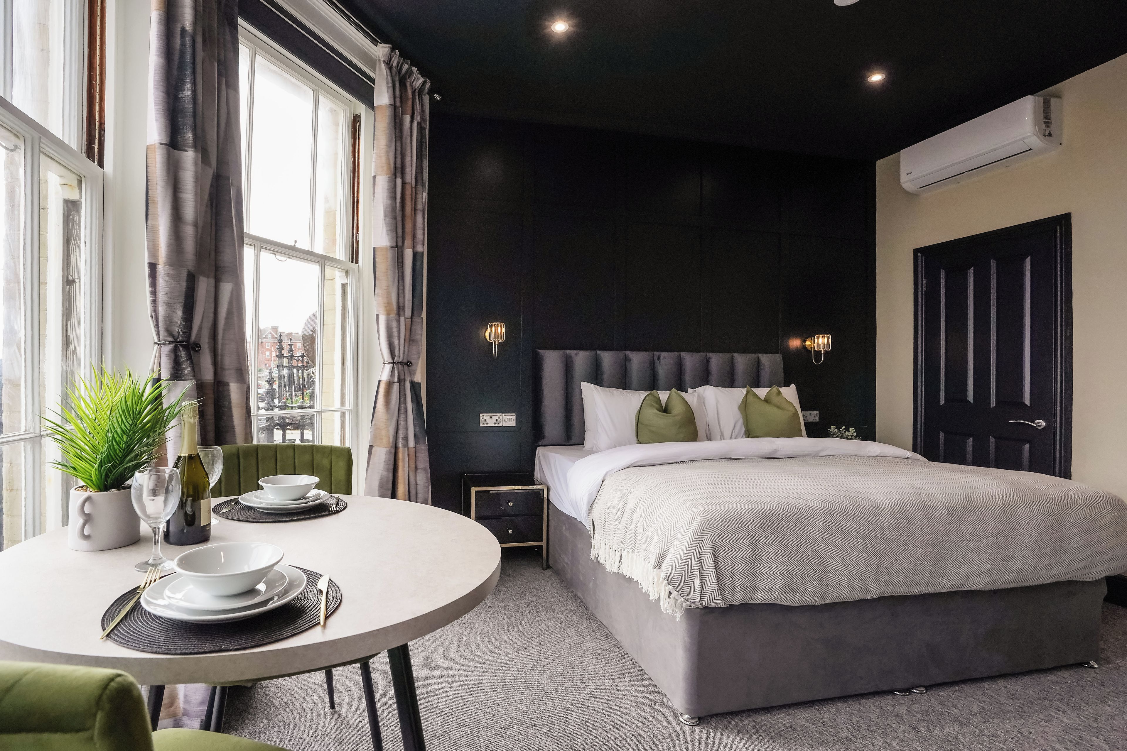 Une chambre d’hôtel moderne avec un grand lit, une table à manger agrémentée d’une plante et une vue sur la ville par la fenêtre.