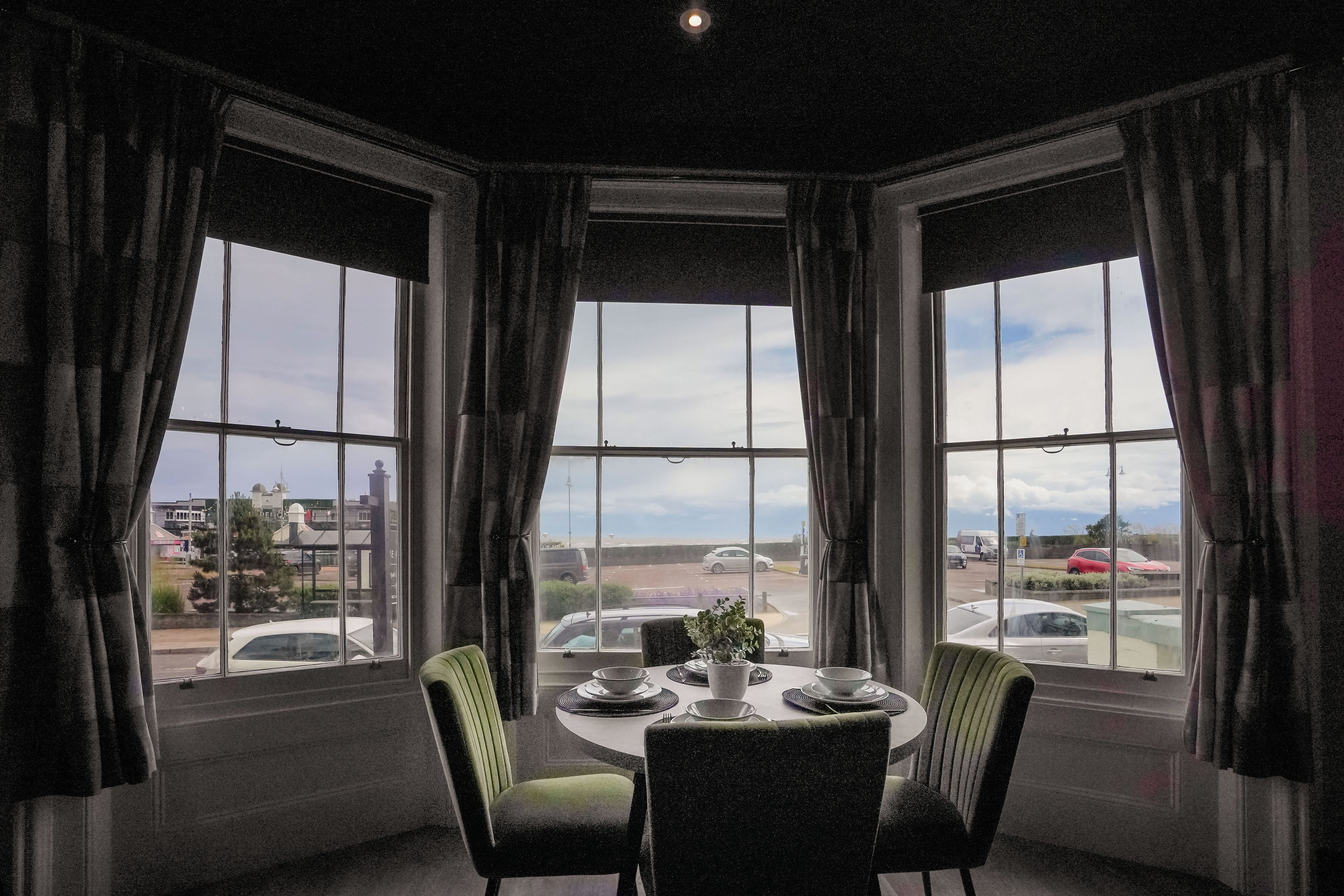 Un comedor con una mesa preparada para cuatro personas, ventanas amplias con cortinas y vista a la calle.