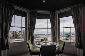 Un comedor con una mesa preparada para cuatro personas, ventanas amplias con cortinas y vista a la calle.