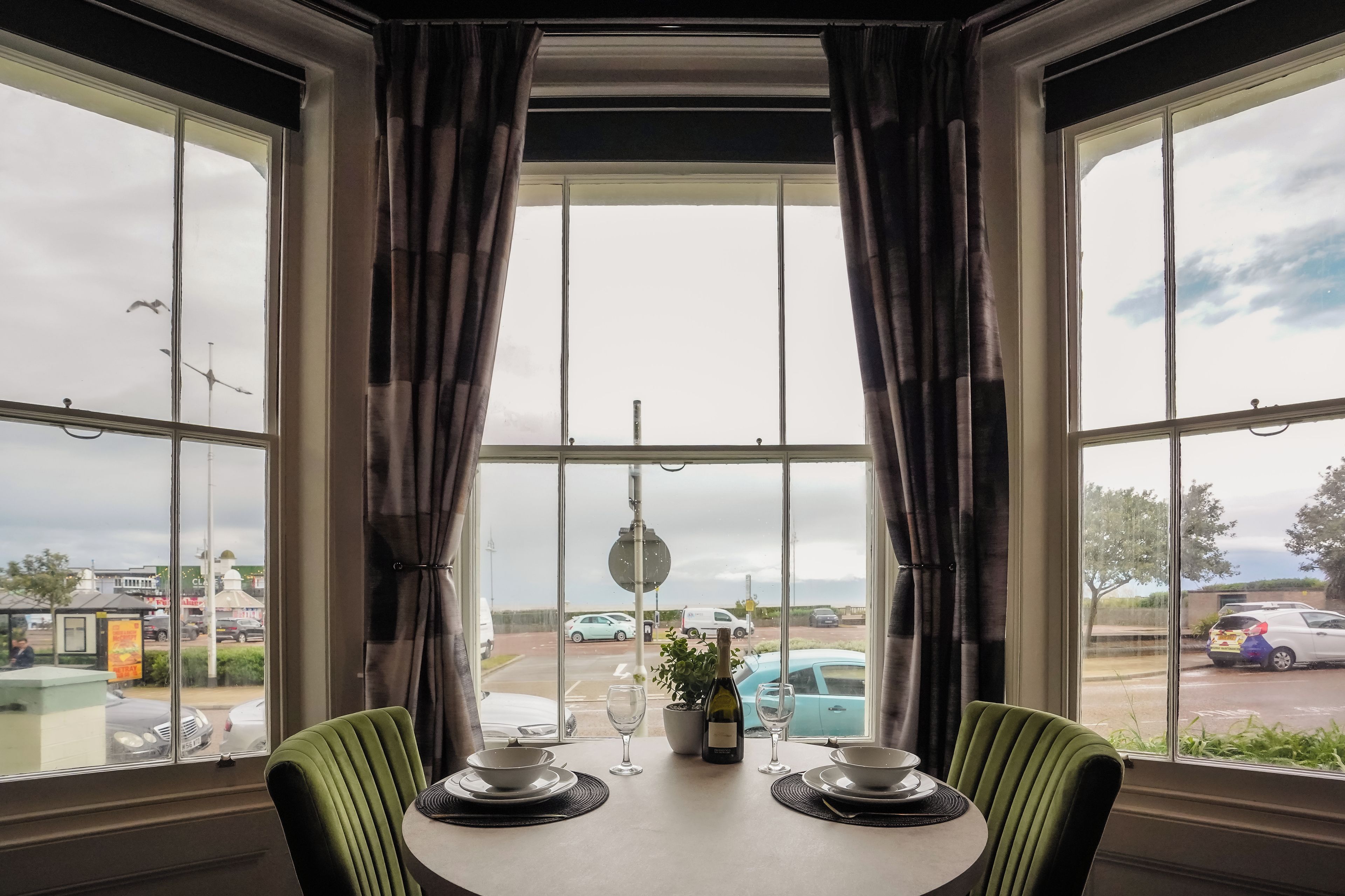 A dining area with a table set for two, large windows with curtains, and a view of a parking lot with cars.