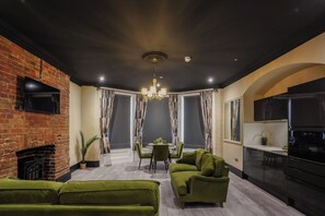 Una sala de estar moderna con sofás verdes, una chimenea, un comedor con una araña y una cocina con estufa negra.