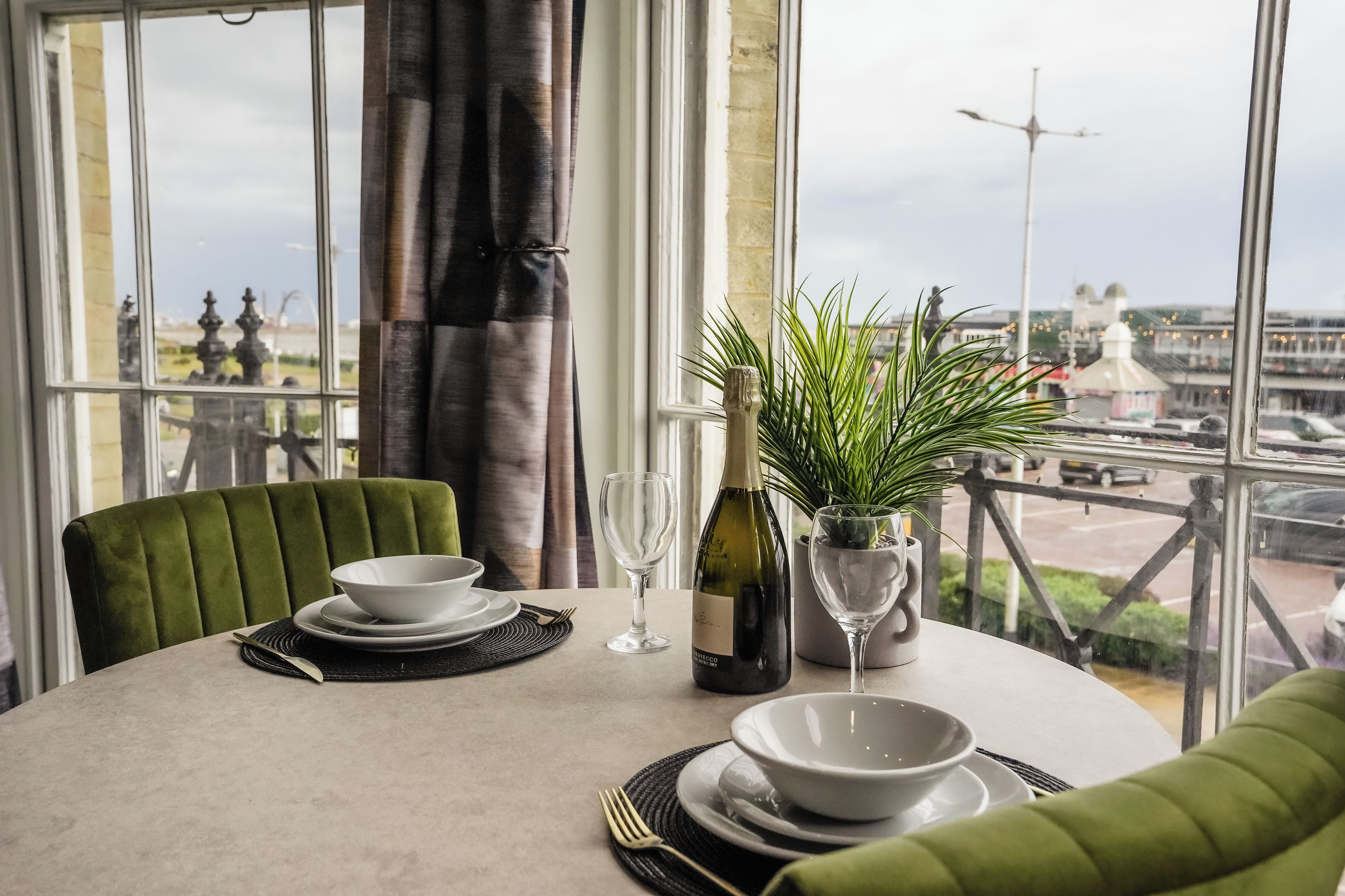 Mesa de jantar posta com pratos, tigelas, garrafa de vinho e um copo, com vista para um horizonte urbano através de grandes janelas.