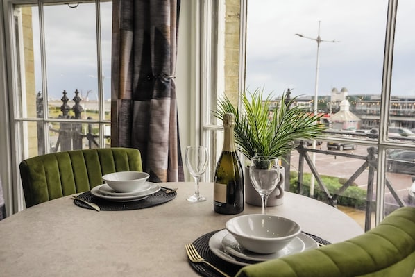 Una mesa de comedor dispuesta con platos, tazones, una botella de vino y una copa, con vistas a un paisaje urbano a través de amplios ventanales.
