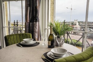 Una mesa de comedor dispuesta con platos, tazones, una botella de vino y una copa, con vistas a un paisaje urbano a través de amplios ventanales.
