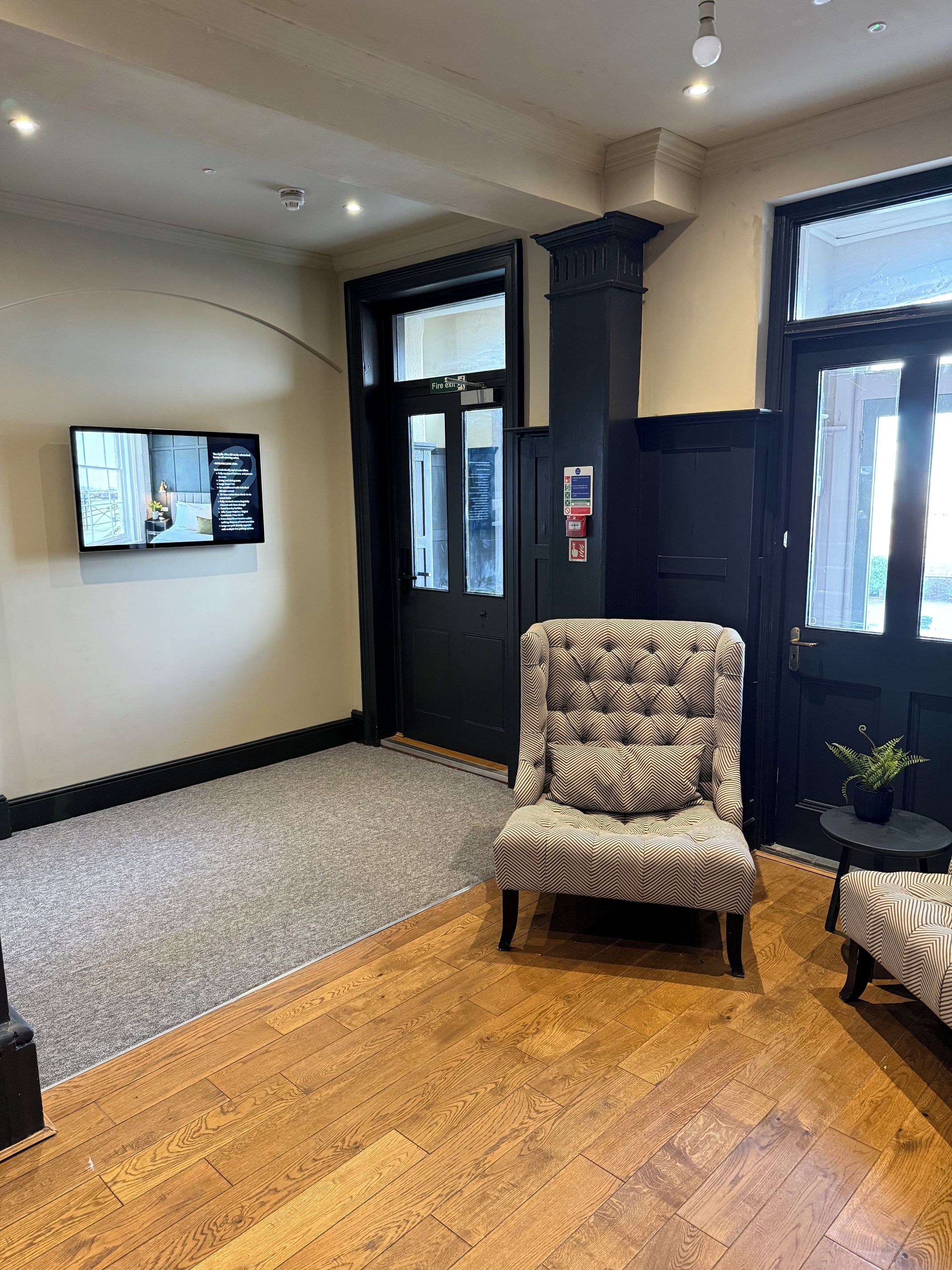 A hotel lobby with a wooden floor, a tufted armchair, a small table with a plant, and a wall-mounted screen displaying information.