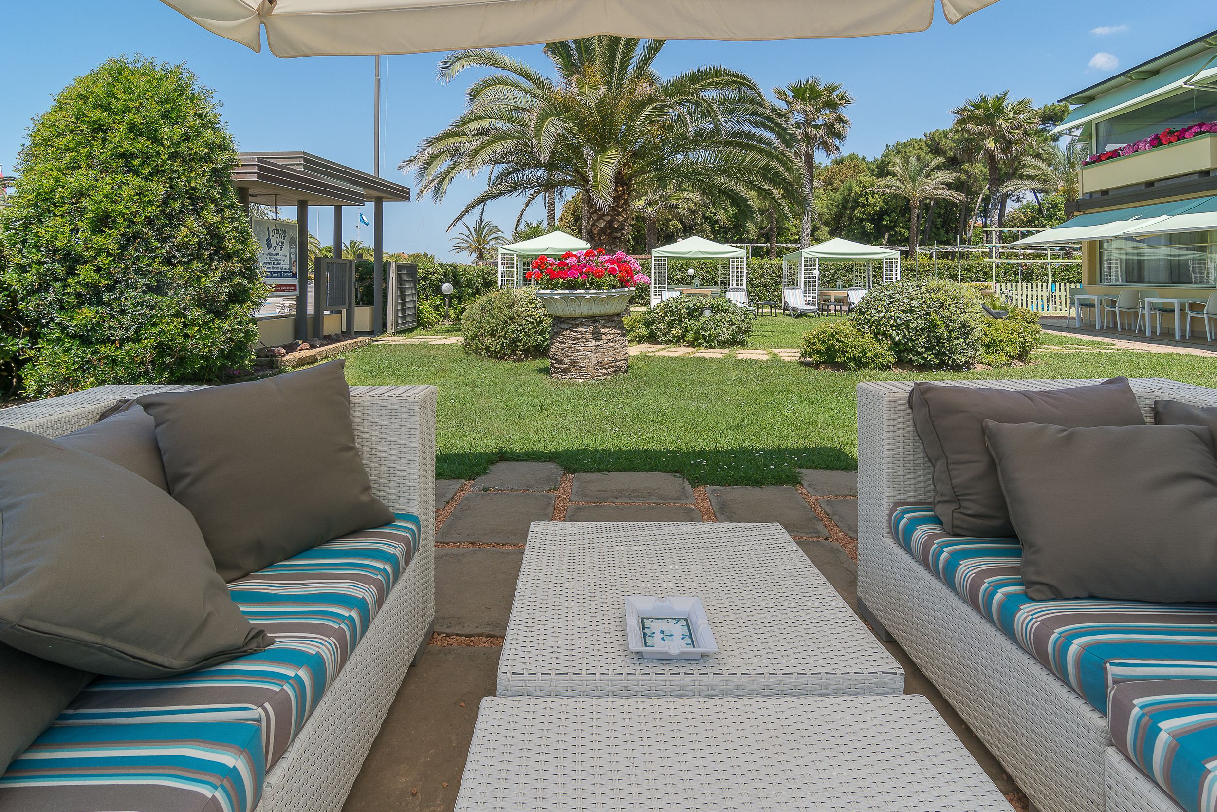 Un patio amueblado con sofás de mimbre, una mesa central y un jardín con palmeras y una fuente.