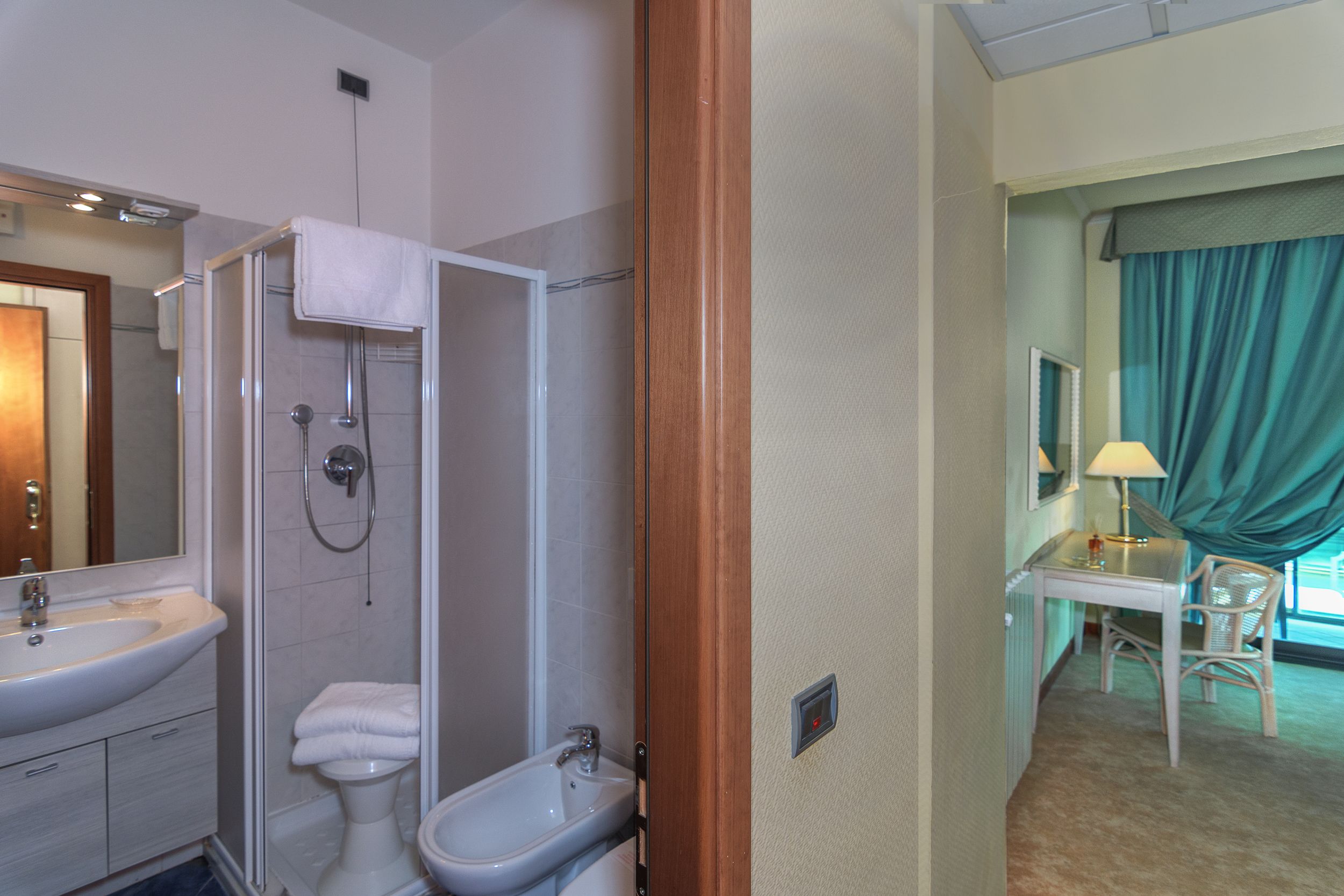 A hotel bathroom with a shower, sink, and bidet. There is a desk and chair in the adjacent room.