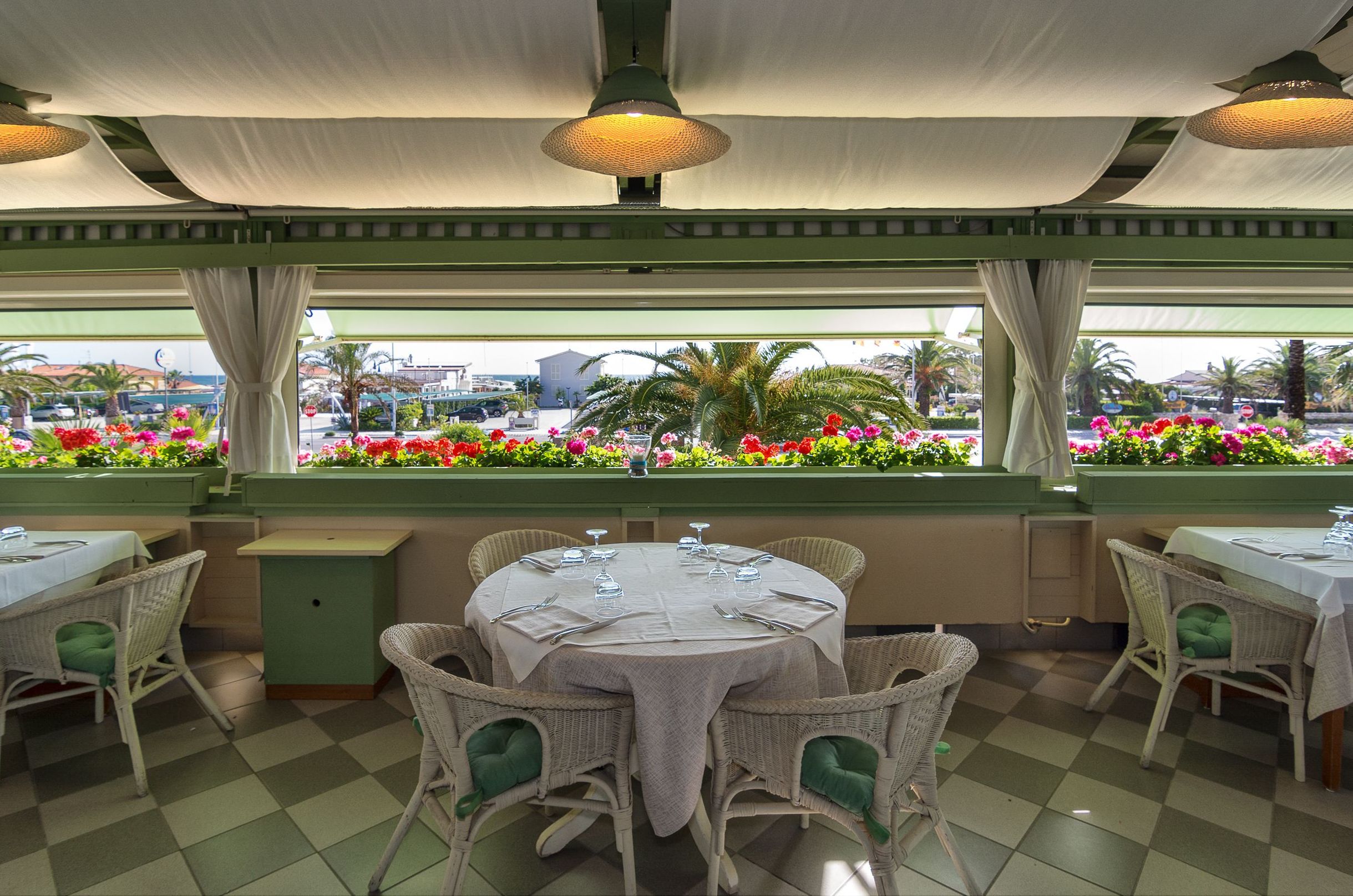 Een restaurant met ronde tafels en rieten stoelen, met uitzicht op de straat met bloemen, en een geruit vloertje.