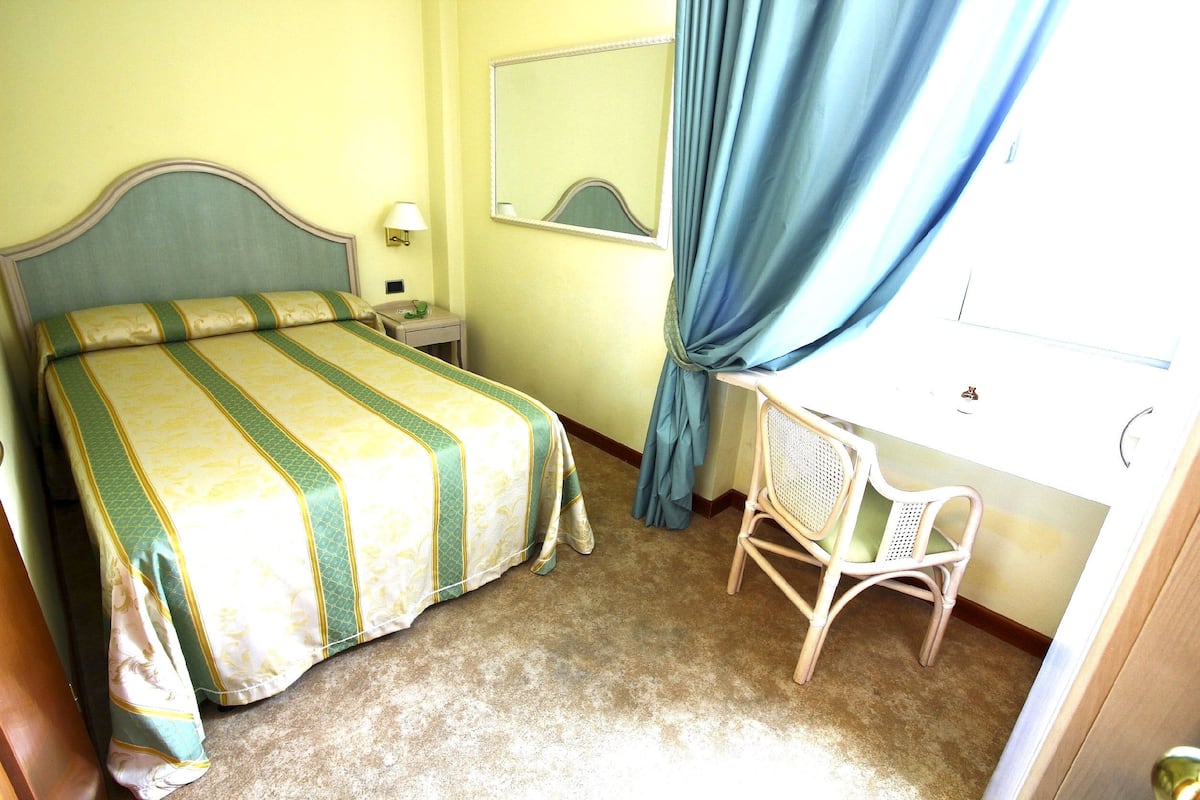 A hotel room with a bed, a nightstand, a chair, a mirror, and a window with curtains.