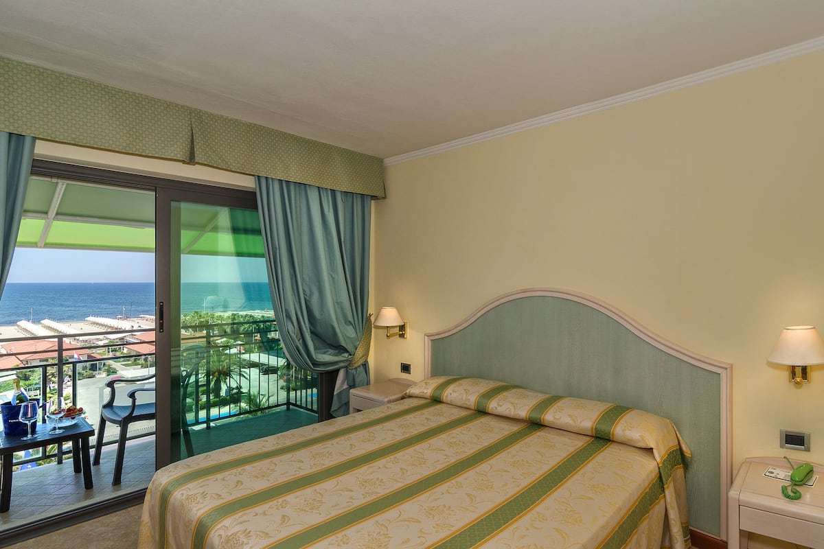 A hotel room with a bed, two bedside lamps, a nightstand, a chair, a table with a drink and glasses, and a view of the ocean through a balcony with curtains.