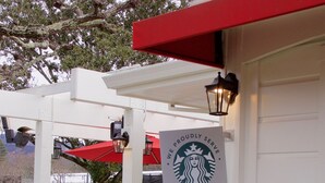 Un espace extérieur avec des tables et des chaises blanches, ainsi qu'un parasol rouge, devant un café Starbucks.
