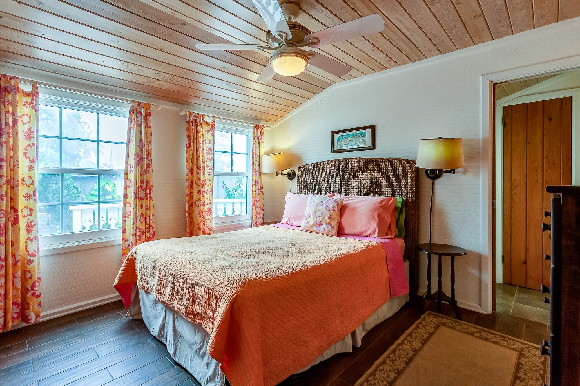 Un dormitorio con techo de madera, una cama con cabecera de mimbre, dos lámparas de noche y una ventana con cortinas florales.