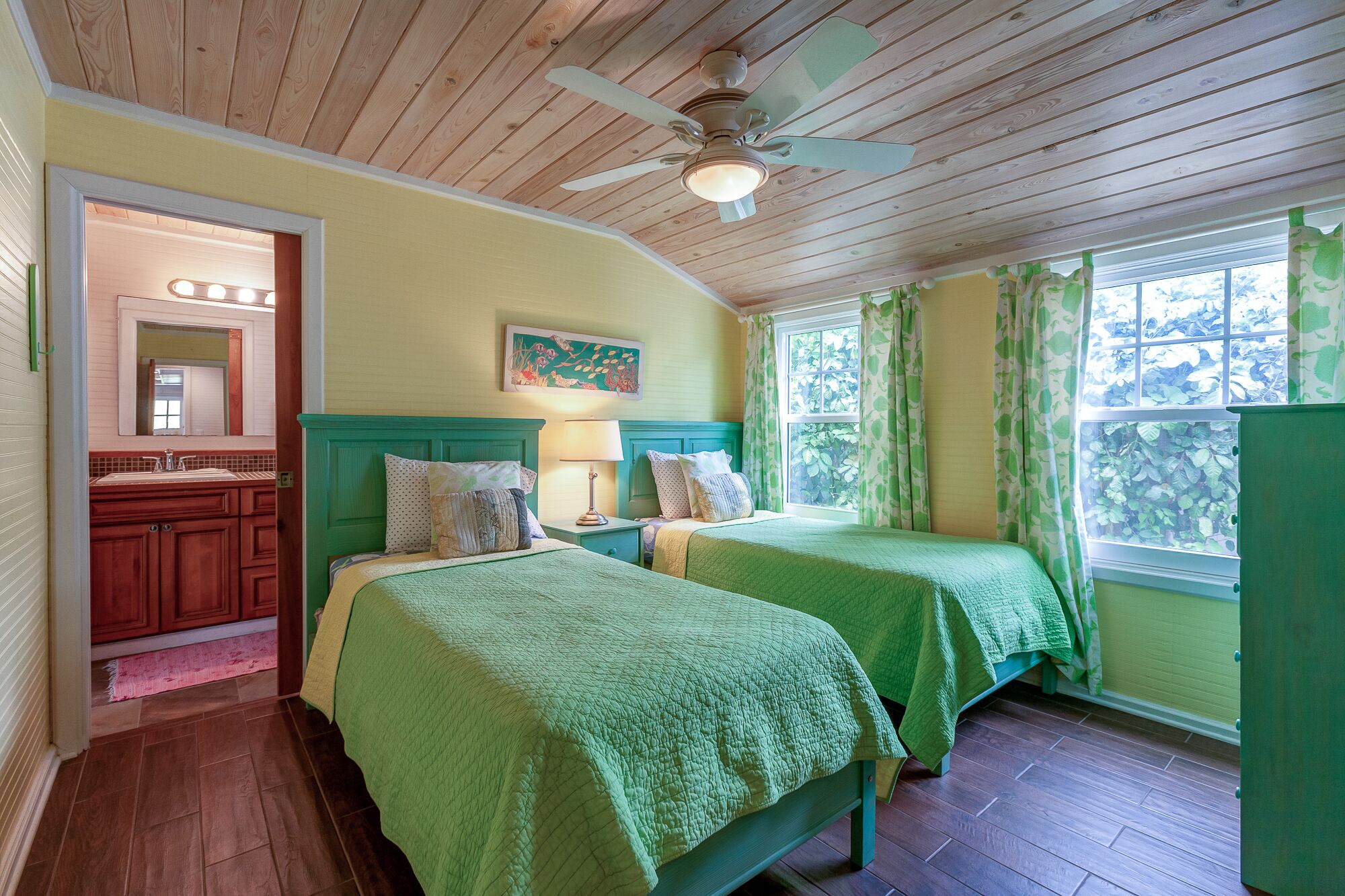 A room with two beds, a wooden ceiling fan, a painting on the wall, and a window with green curtains.