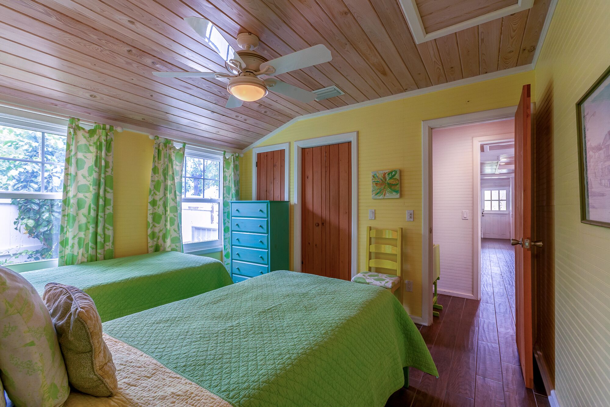 A room with two beds, a ceiling fan, wooden walls, a window with curtains, and a door leading to another room.