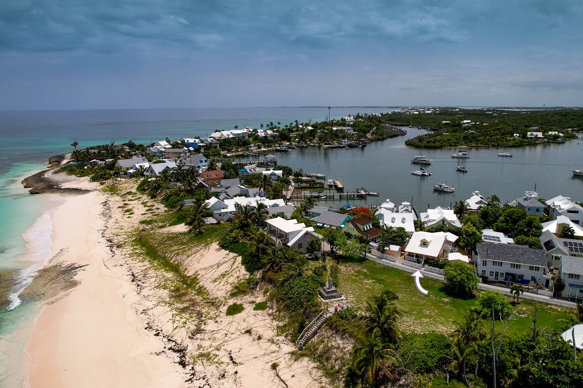 Vista aérea de un pueblo costero con casas a lo largo de la playa, un puerto deportivo con barcos y una vista despejada del océano.