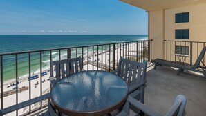 A balcony with a glass table and chairs overlooking a beach with umbrellas and lounge chairs.