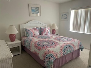 A bedroom with a white bed, patterned bedding, two bedside lamps, a wicker chair, and a window with blinds. - Shorewalk 205 - Bradenton-  The Palms condo 2 Bedroom / 2 Bath,  5 miles from  Anna Maria Island and near Img, maximum occupancy of 4 people. (Bradenton)